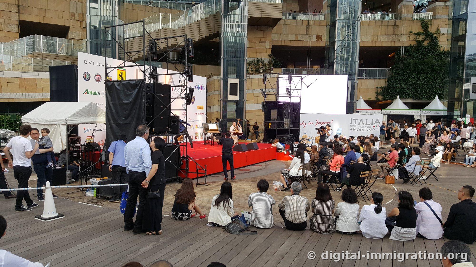 Événement culturel italien sur la scène extérieure de l'arène de Roppongi Hills avec un musicien en kimono sur scène rouge et un décor 'ITALIA AMORE mio', des spectateurs assis sur une terrasse en bois