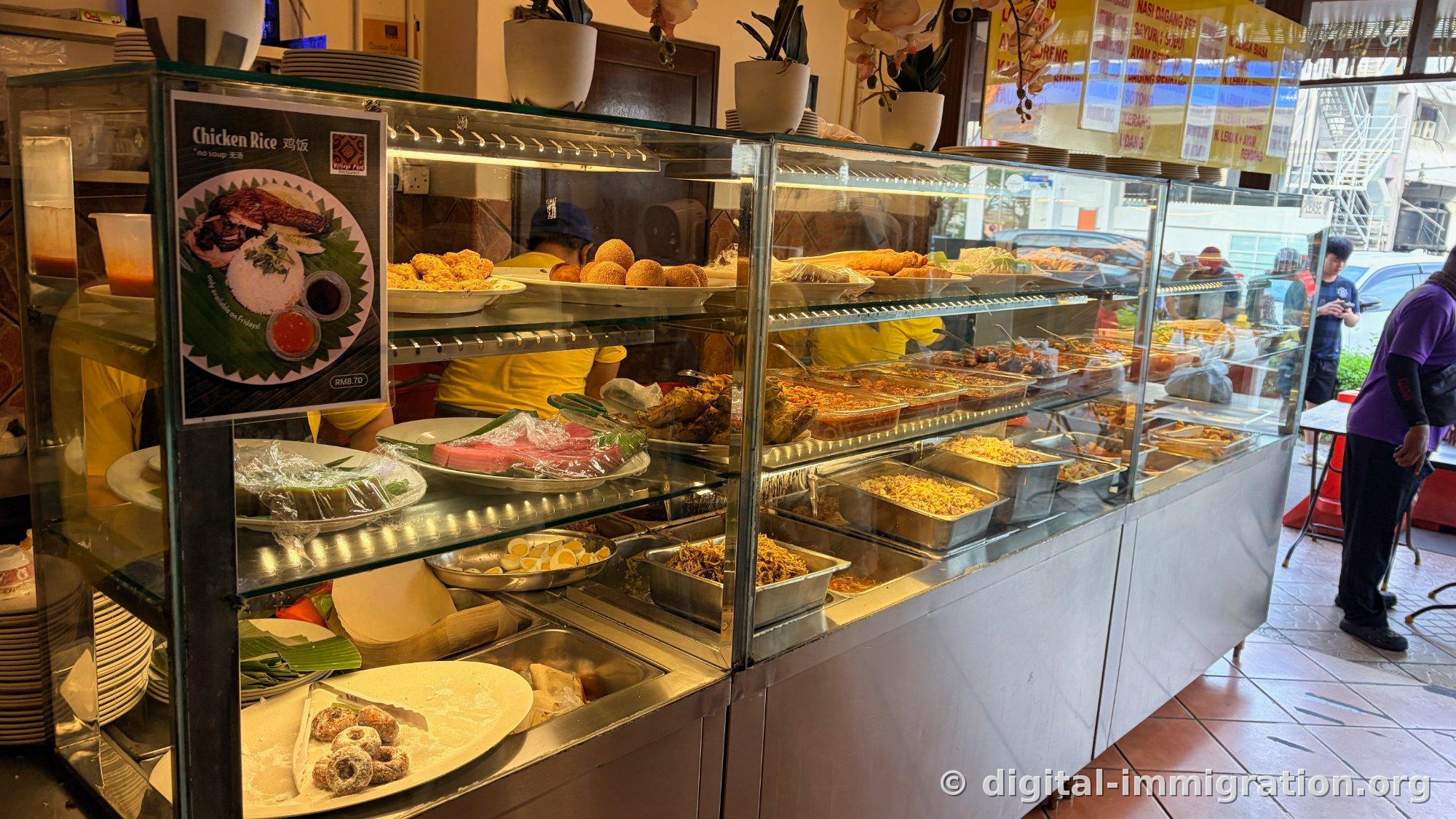 Inside a glass display case, sesame fried bread, sugar donuts, boiled eggs, fried anchovies, and skewers are stacked in layers, with a RM8.70 Chicken Rice sign on the left inside the store