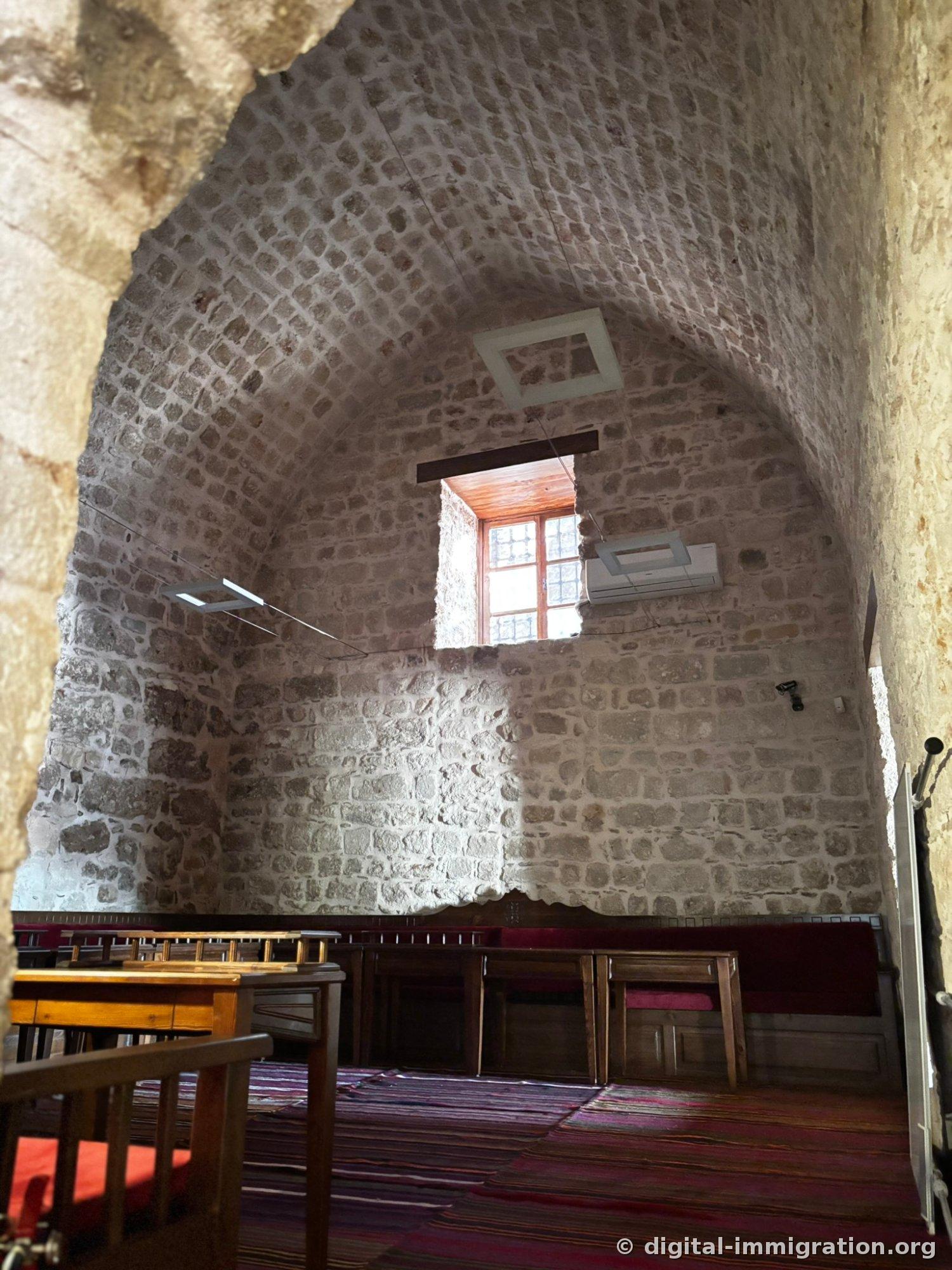 Salle au haut plafond voûté dont le plâtre s'est écaillé révélant la pierre, un rayon de lumière carré d'une fenêtre, bancs en bois et coussin mauve sous une carpette rayée