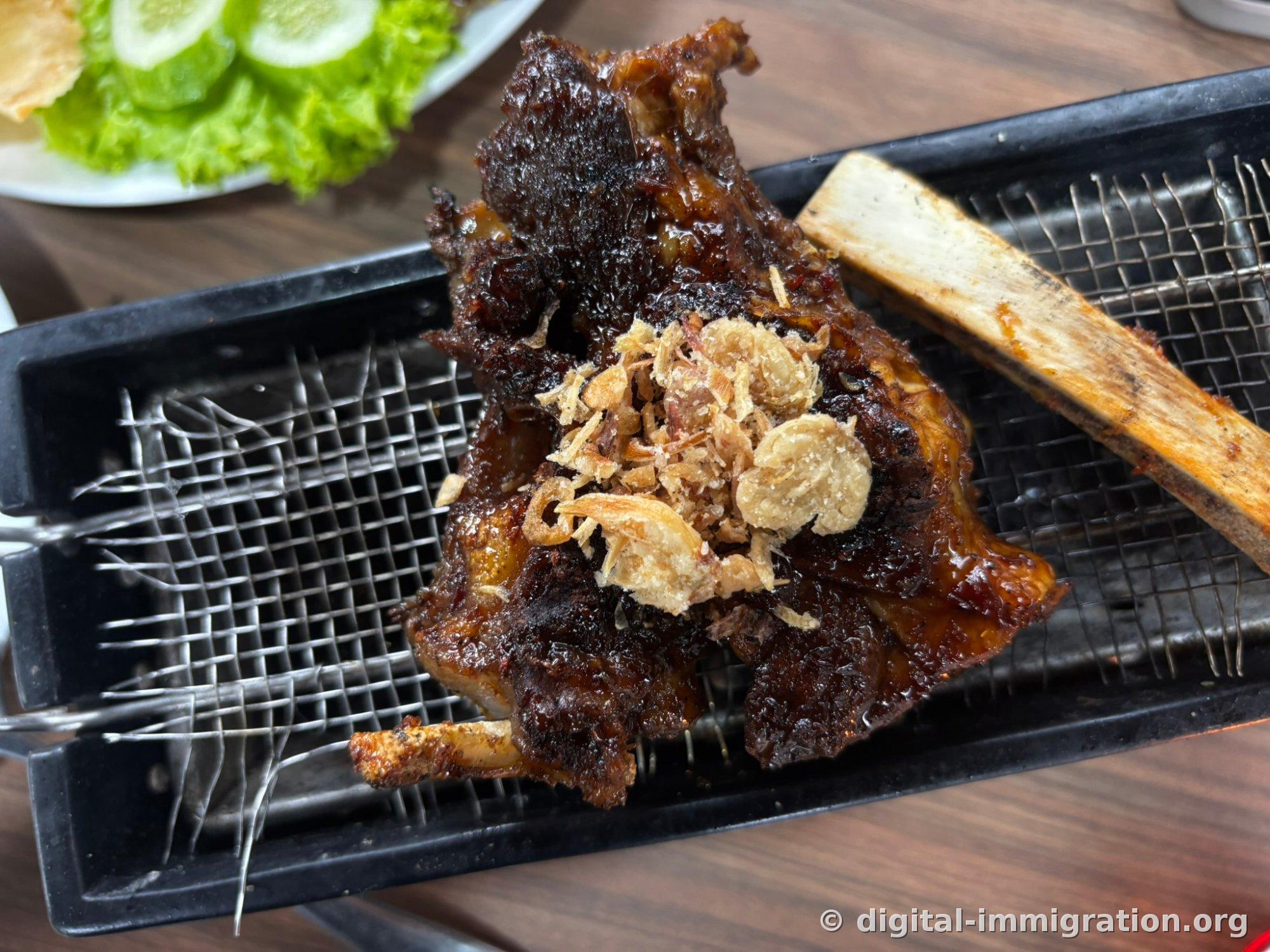 Soy-glazed grilled beef short ribs placed on a black wire grill, heaped with fried shallots on top, with lettuce and cucumber on a side plate