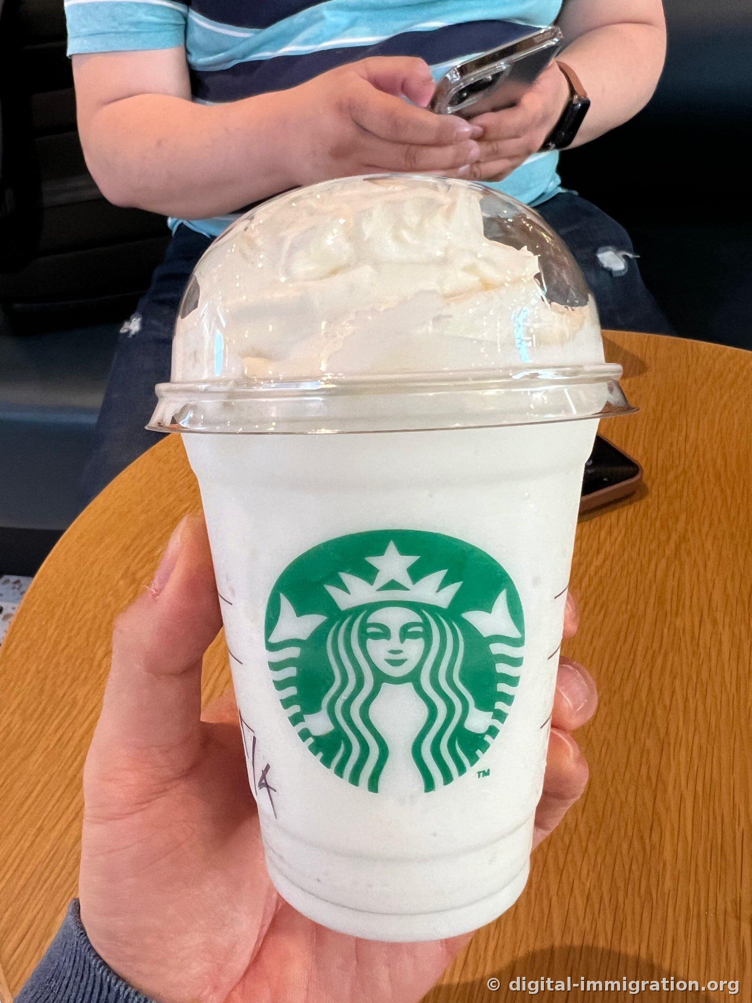 Une boisson frappuccino blanc Starbucks tenue à la main, avec de la crème fouettée remplissant généreusement le couvercle en dôme, et des compagnons assis derrière regardant leur téléphone