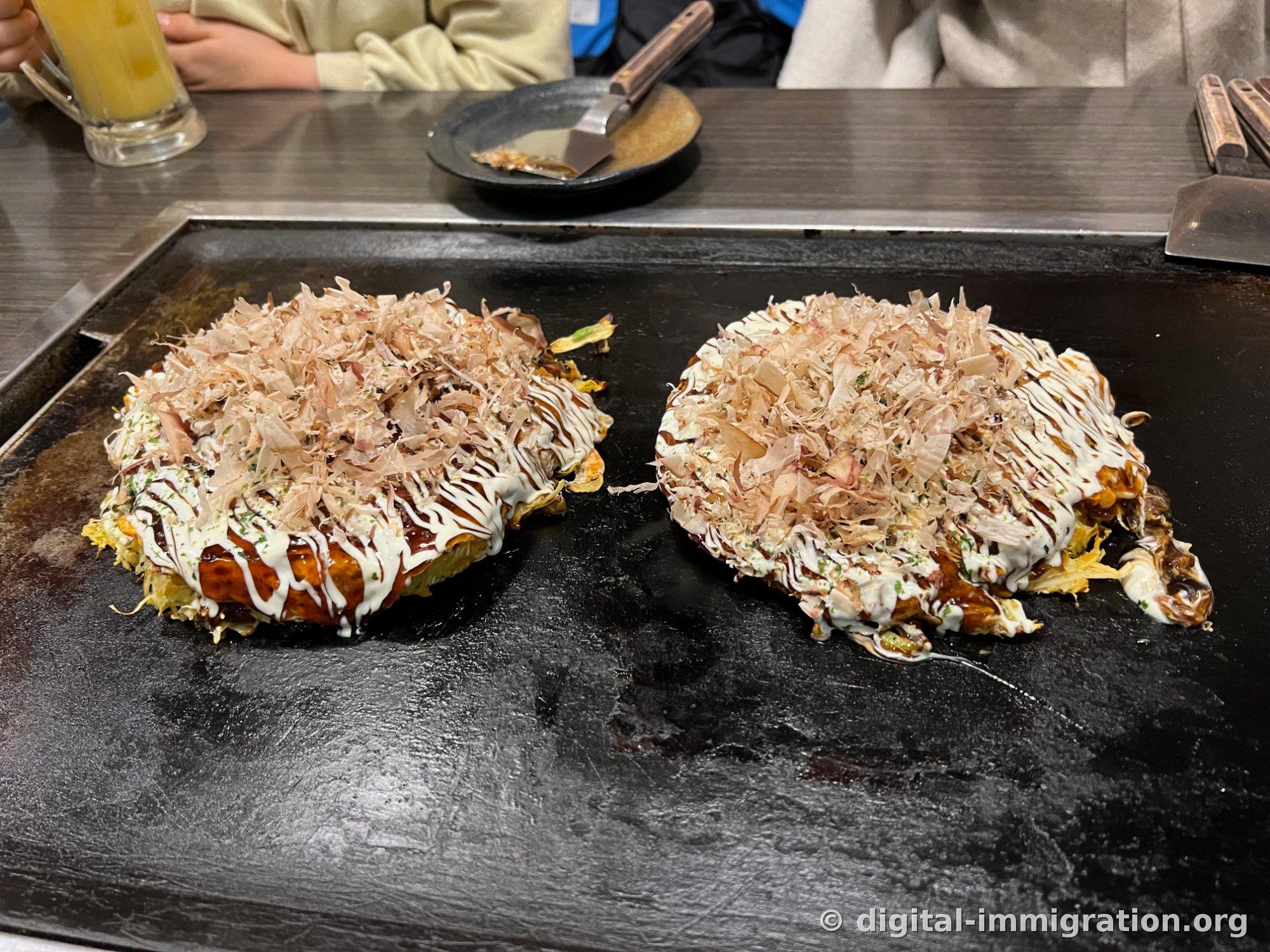 Deux okonomiyaki alignés sur la plaque, avec de la sauce et de la mayonnaise versées, généreusement couverts de bonite séchée
