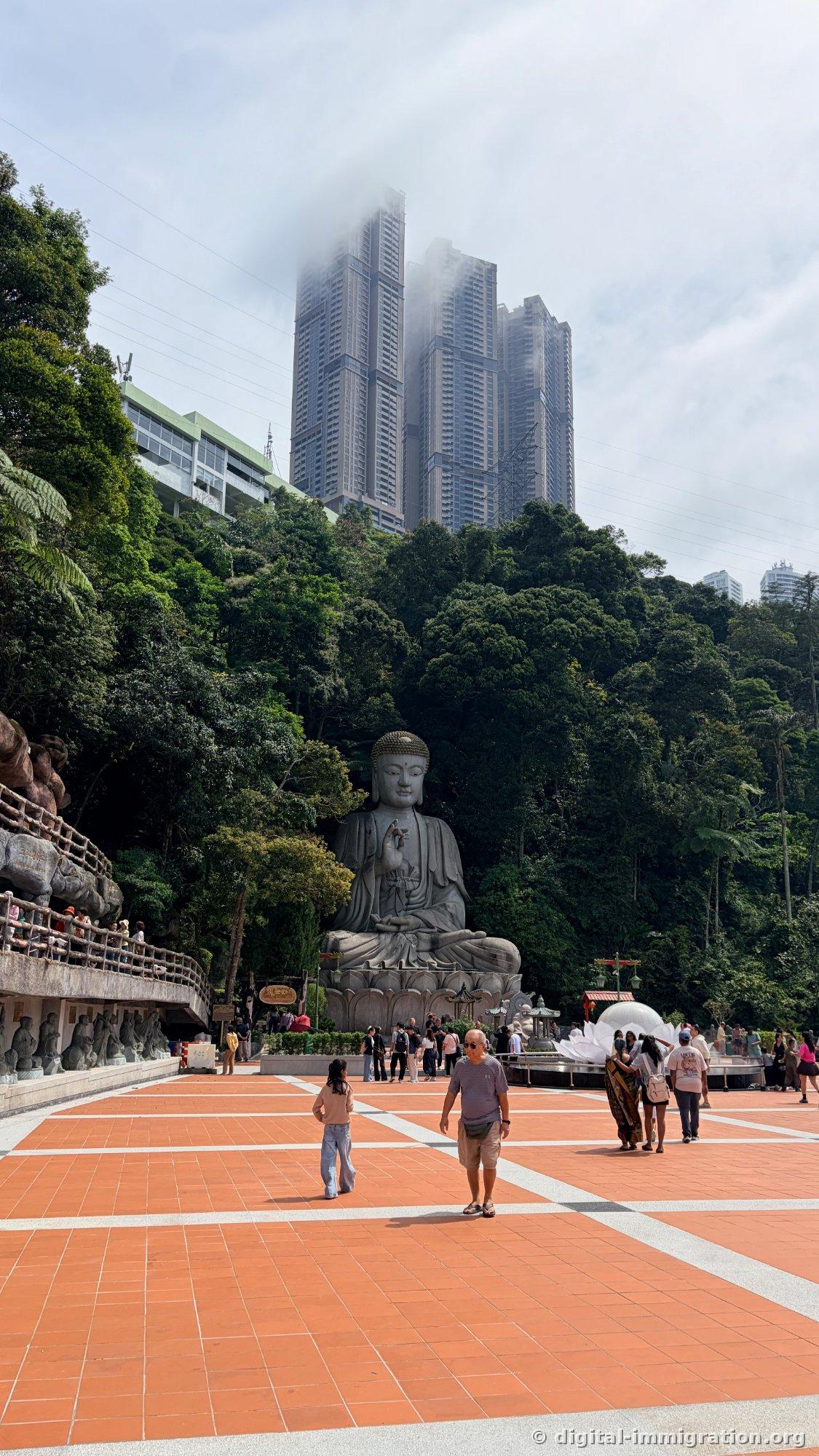 Une gigantesque statue assise en pierre de Bouddha sur une large place en carrelage orange, avec une forêt en arrière-plan et une tour de condominium de grande hauteur enveloppée de nuages au-dessus