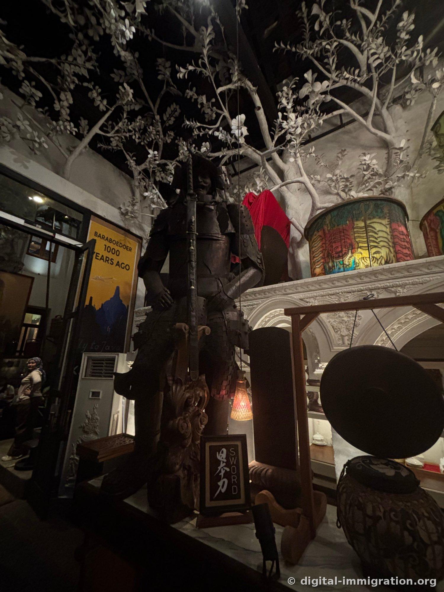 Armored samurai statue with sword beneath concrete tree, next to old BARABOEDOER 1000 YEARS AGO travel poster and gong
