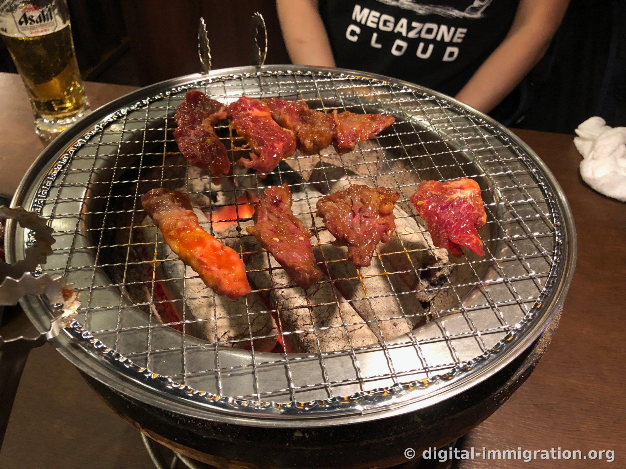 Eight pieces of marinated meat on the shichirin grate with red flames visible between coals, an Asahi draft beer glass and tongs beside it