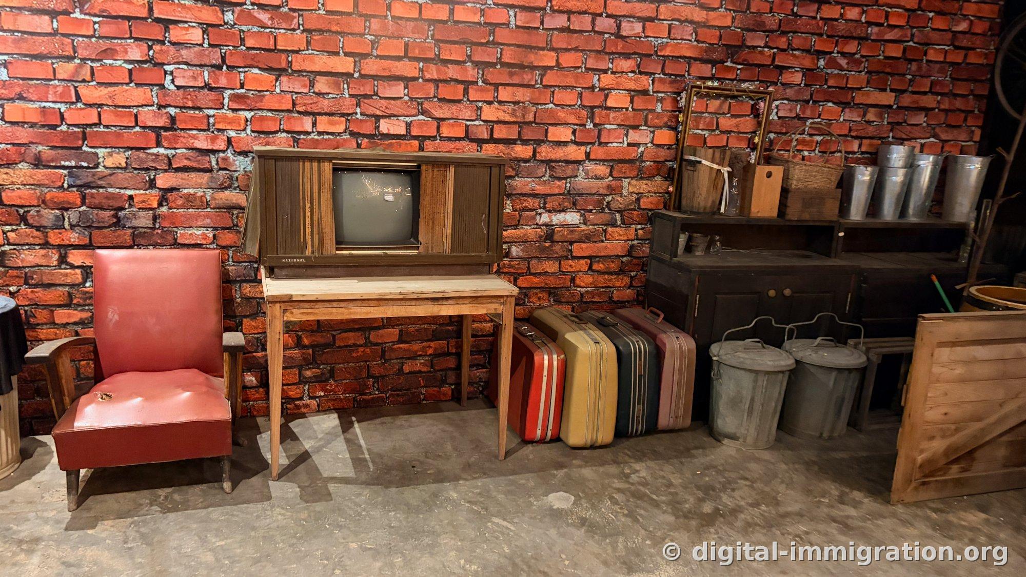 Devant un mur de brique rouge, une télévision NATIONAL vintage avec meuble en bois, un fauteuil en simili-cuir rouge, des valises de voyage vintage aux couleurs variées et des poubelles en tôle