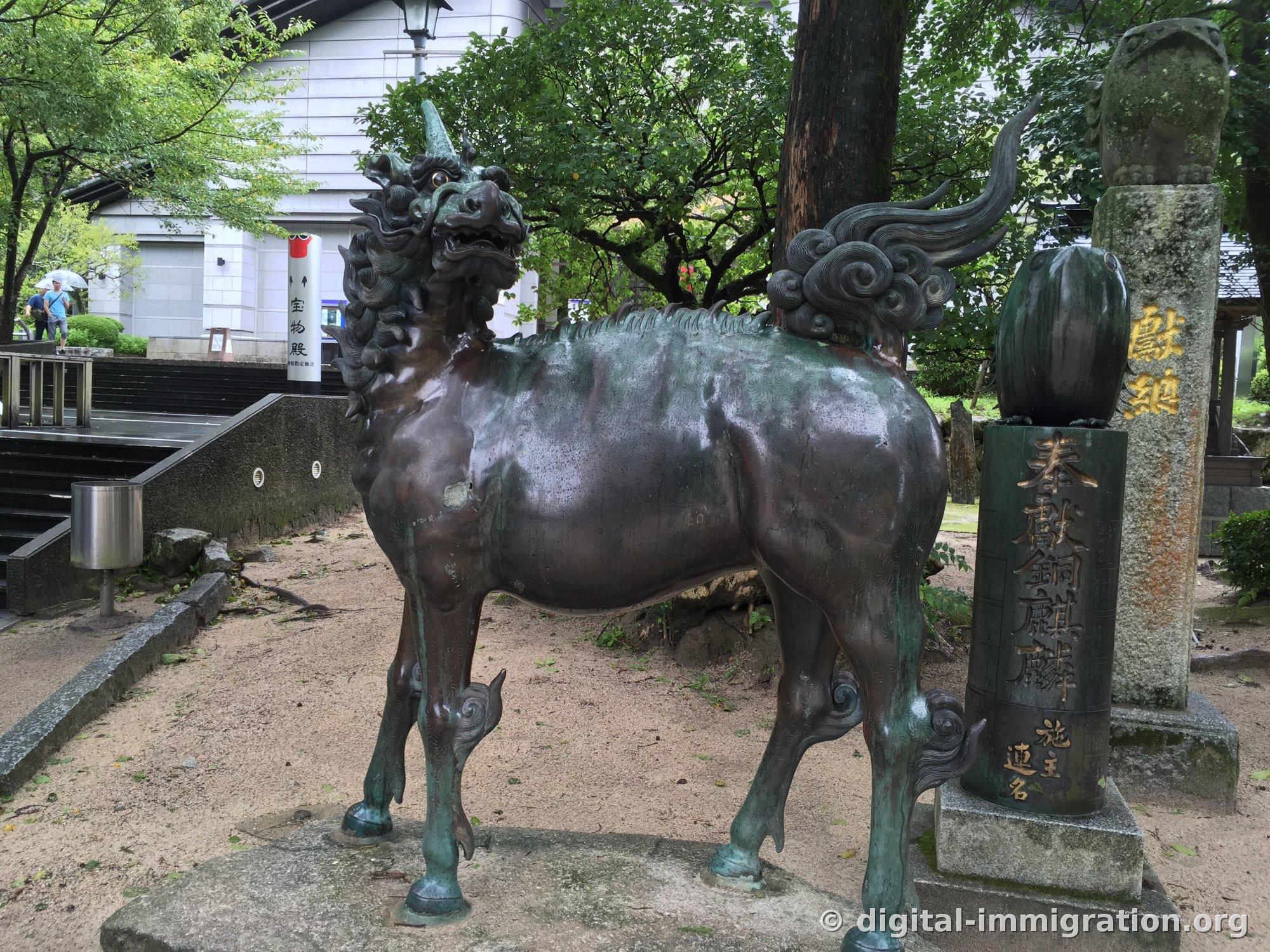 Bronze unicorn statue standing next to a stone marker inscribed '奉献 銅麒麟', treasure house building in the background