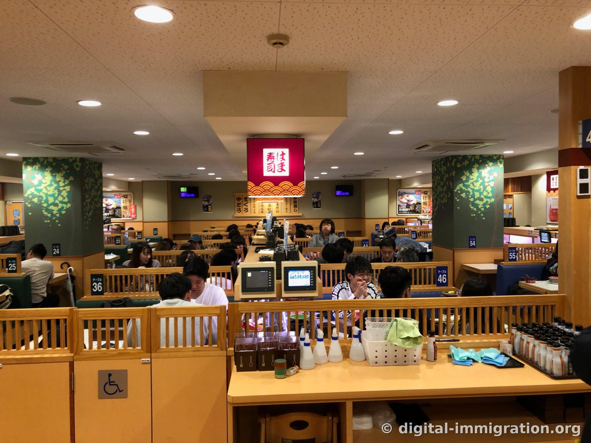 Une lanterne Hamasushi rouge est suspendue au plafond d'une grande salle avec des numéros de table 24, 36, 46, 54 et des box où les clients sont assis