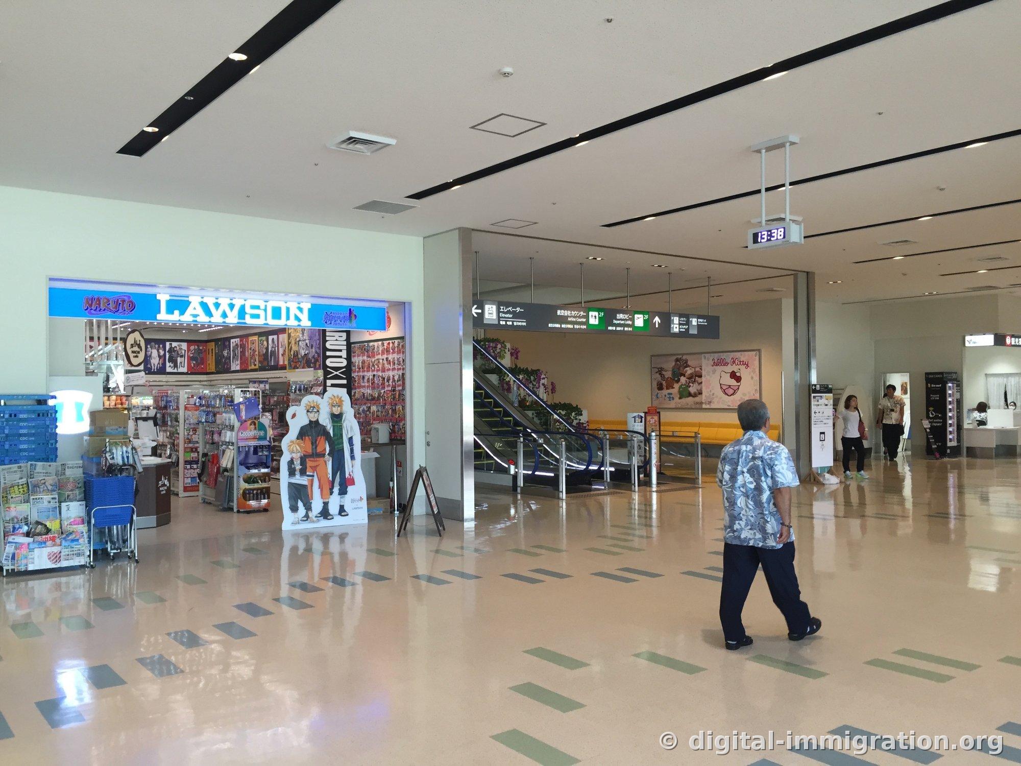 Entrée du magasin Lawson au 1er étage du terminal des vols domestiques de l'aéroport de Naha. Il y a une pancarte de collaboration Naruto et des panneaux de taille réelle, et à droite on voit l'escalier roulant pour monter au hall de départ du 2e étage et les panneaux d'information.