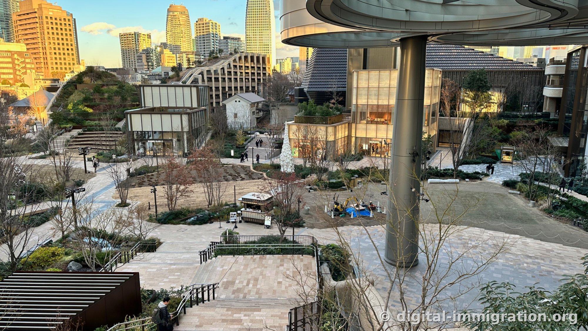 View of Azabudai Hills' central plaza from above the stairs, with wooden kiosk and operating excavator, Hermès store glass facade on the right