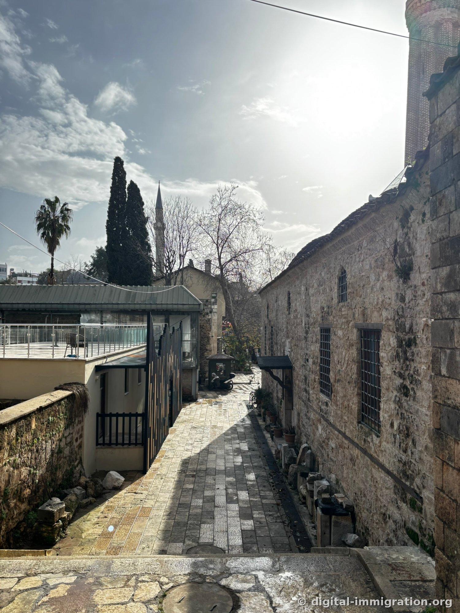 A narrow stone-paved alley. On the right, an ancient limestone wall with iron-barred windows; on the left, a modern glass and black metal cafe terrace; a minaret spire and cypress tree visible in the distance.