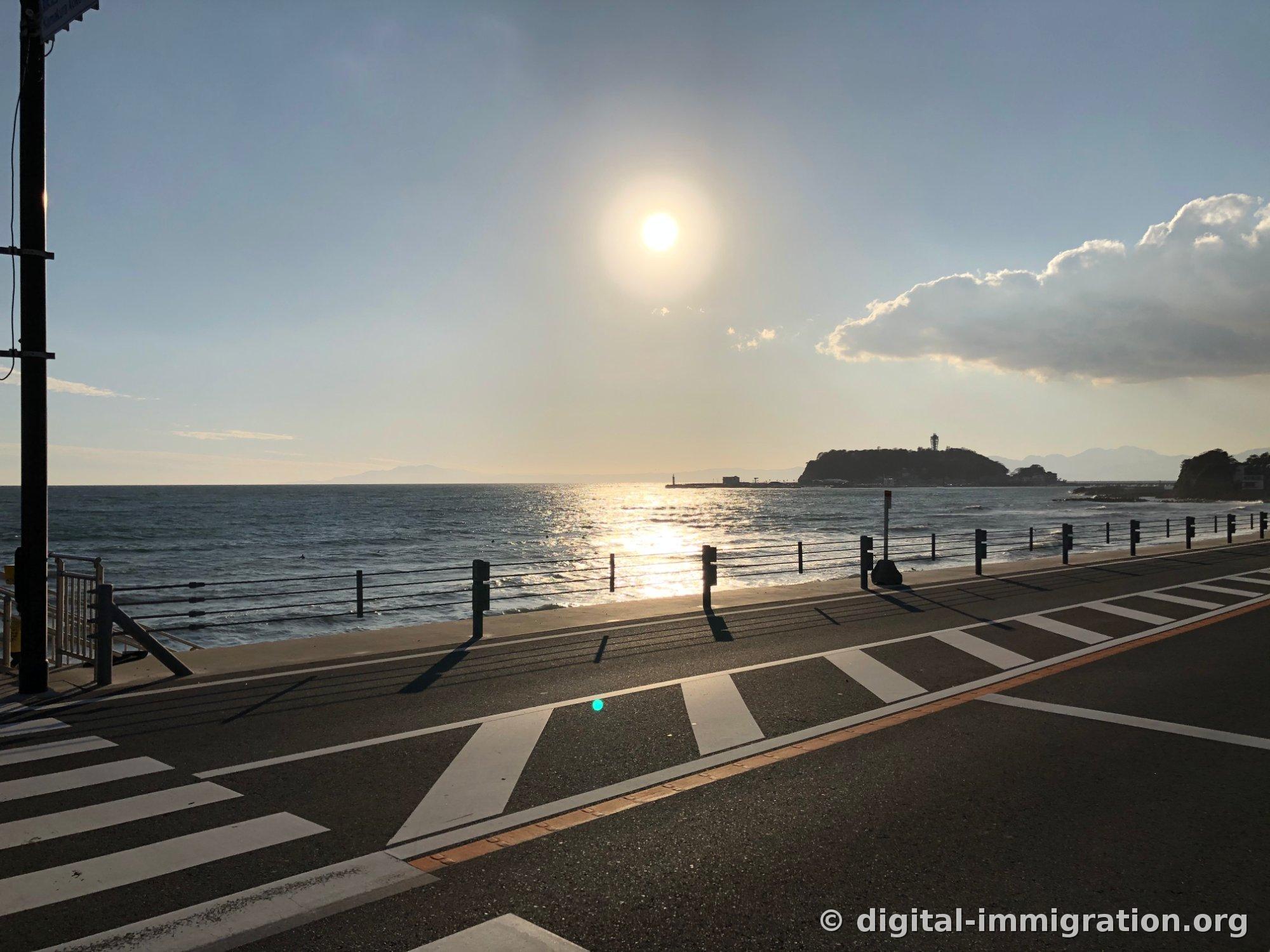 Au-dessus de la mer avec le soleil bas, une colonne de lumière s'étend, l'île d'Enoshima et l'observatoire en silhouette au loin à droite, scène de passage clouté au bord de la route