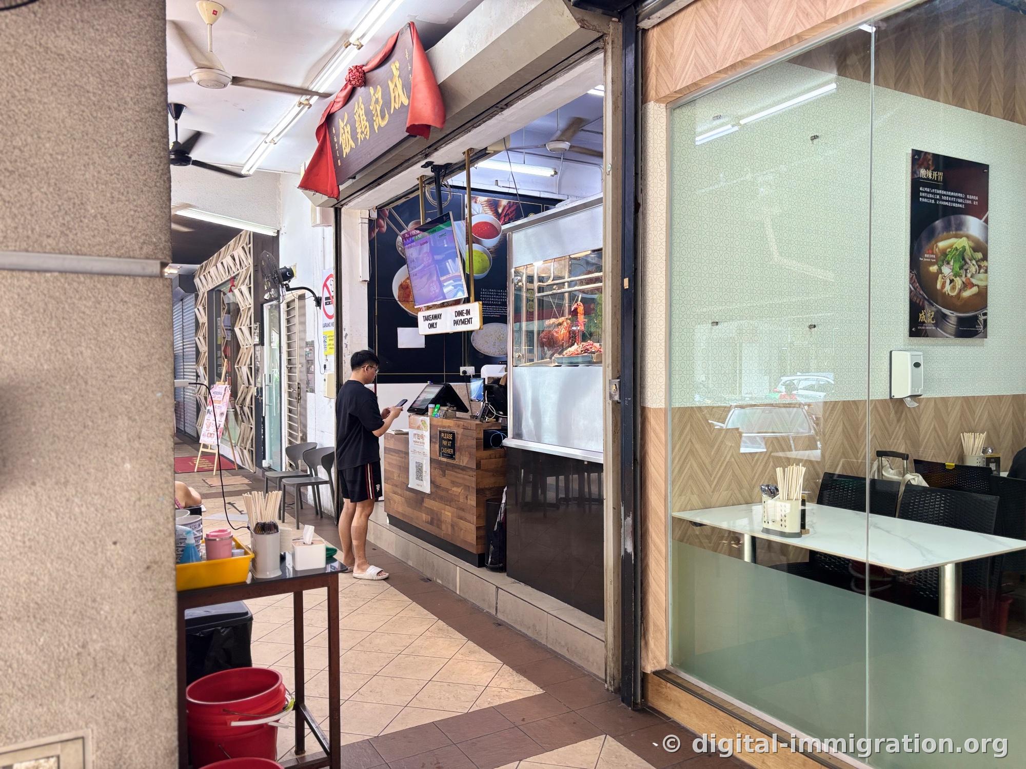 The store entrance with a 成記雞飯 plaque draped in red fabric. A customer stands at the counter checking their phone while waiting to order, with a roasted chicken display case on the right
