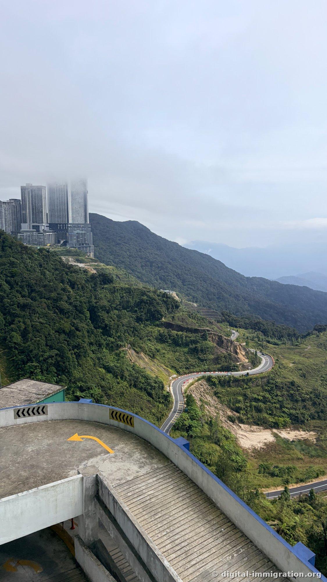 Cloudy sky above mountain ridge with high-rise apartments and winding road circling mountainside, with yellow arrow on parking ramp in foreground