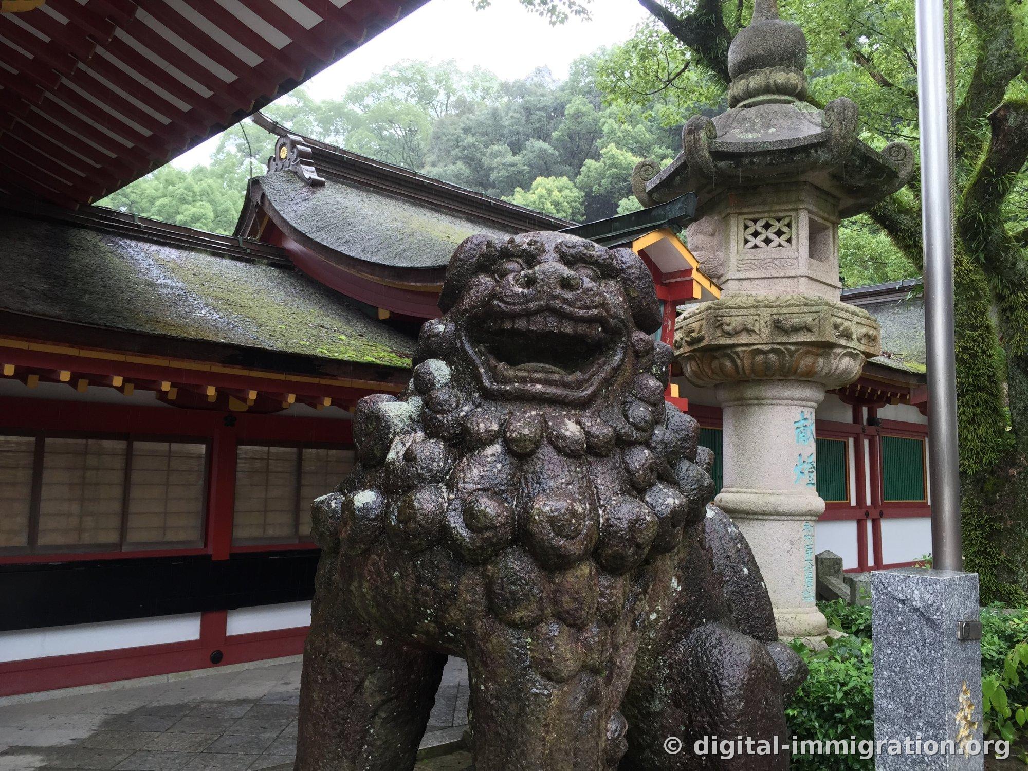 Stone komainu statue with open mouth in front of the corridor and stone lantern beyond, corridor building with red pillars