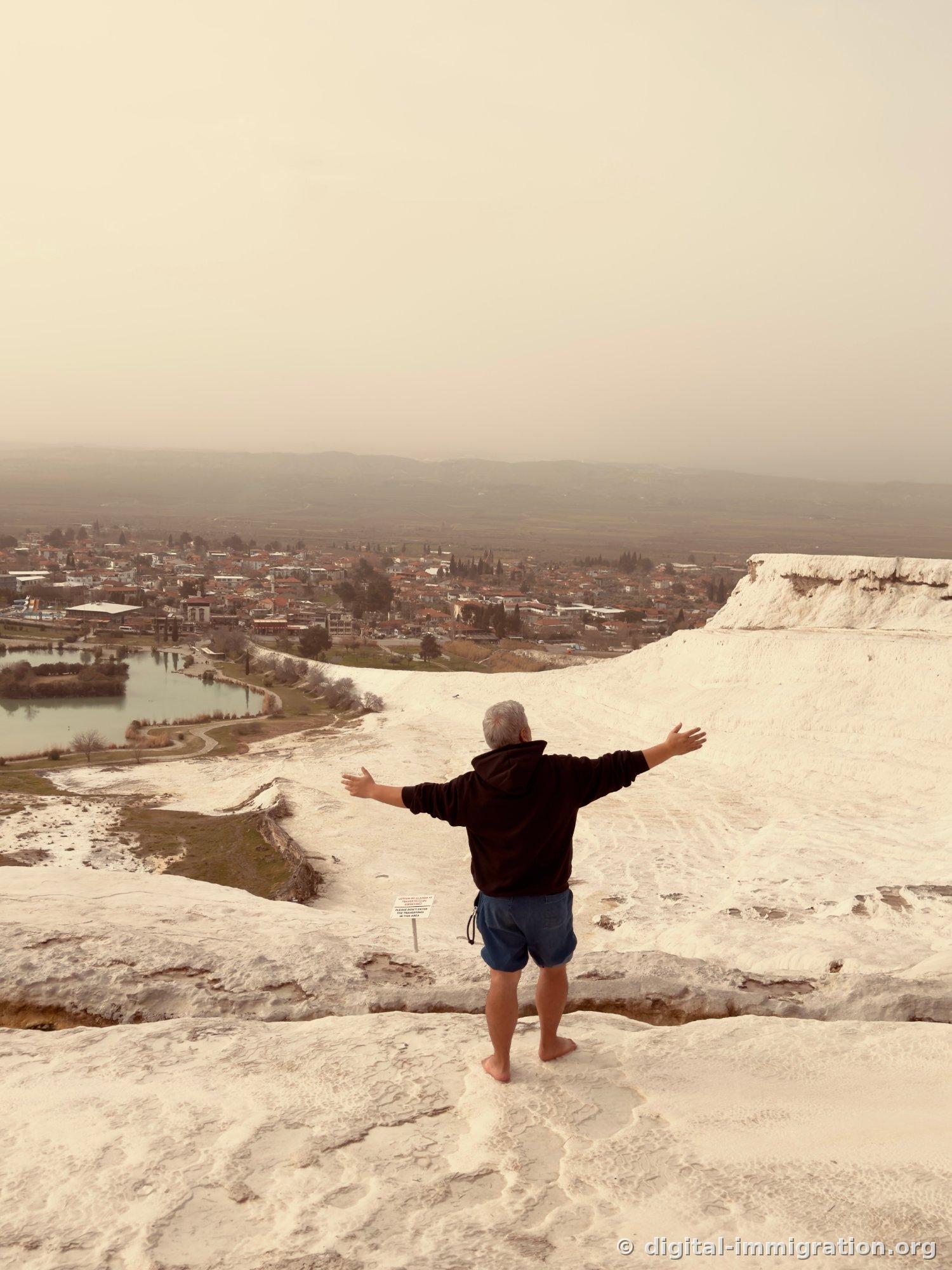 Back view of a person standing on a white limestone ridge with arms spread wide toward the village, with a green lake and the village of Pamukkale visible on the lower left