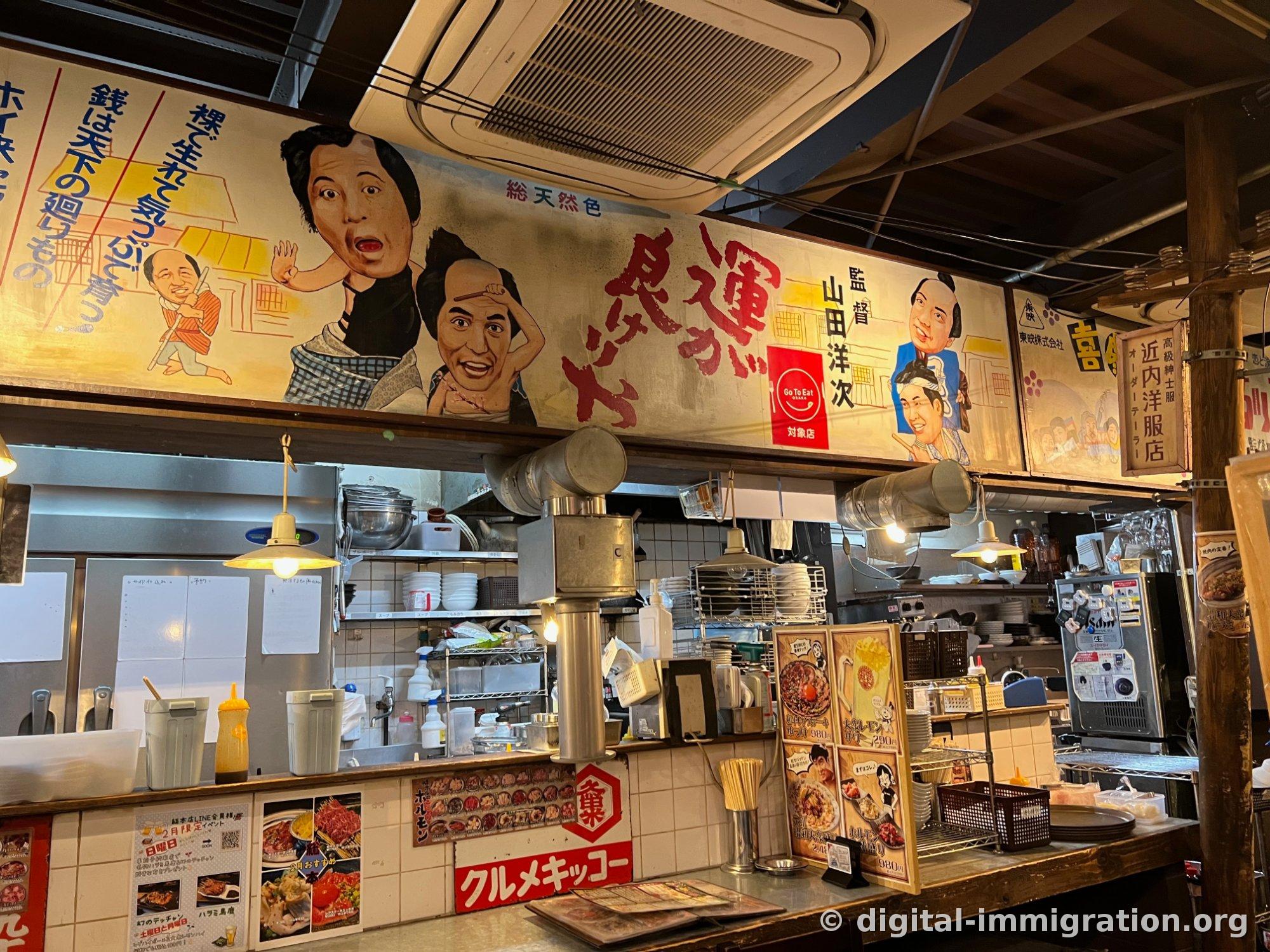 昭和時代の映画看板がかかった店のカウンター。手描きの俳優の顔と縦の日本語の文句、下の方はオープンキッチンとメニュー写真、赤い看板が見える