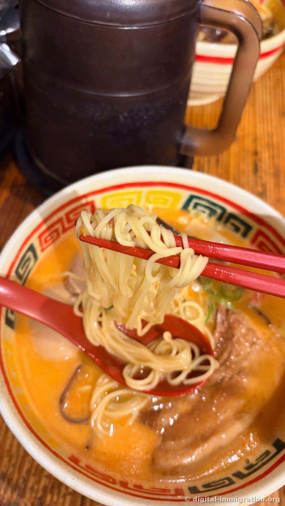 Thin straight noodles lifted with red chopsticks, the bowl below showing yellow tonkotsu broth with kakuni and scallion, with a water bottle and another bowl visible in the background