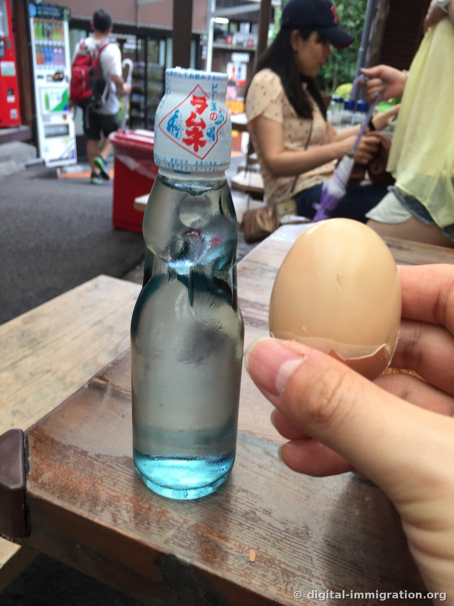 Une bouteille de ramune avec des billes sur une table en bois et un œuf onsen décortiqué dans la main, avec la boutique et les distributeurs en arrière-plan