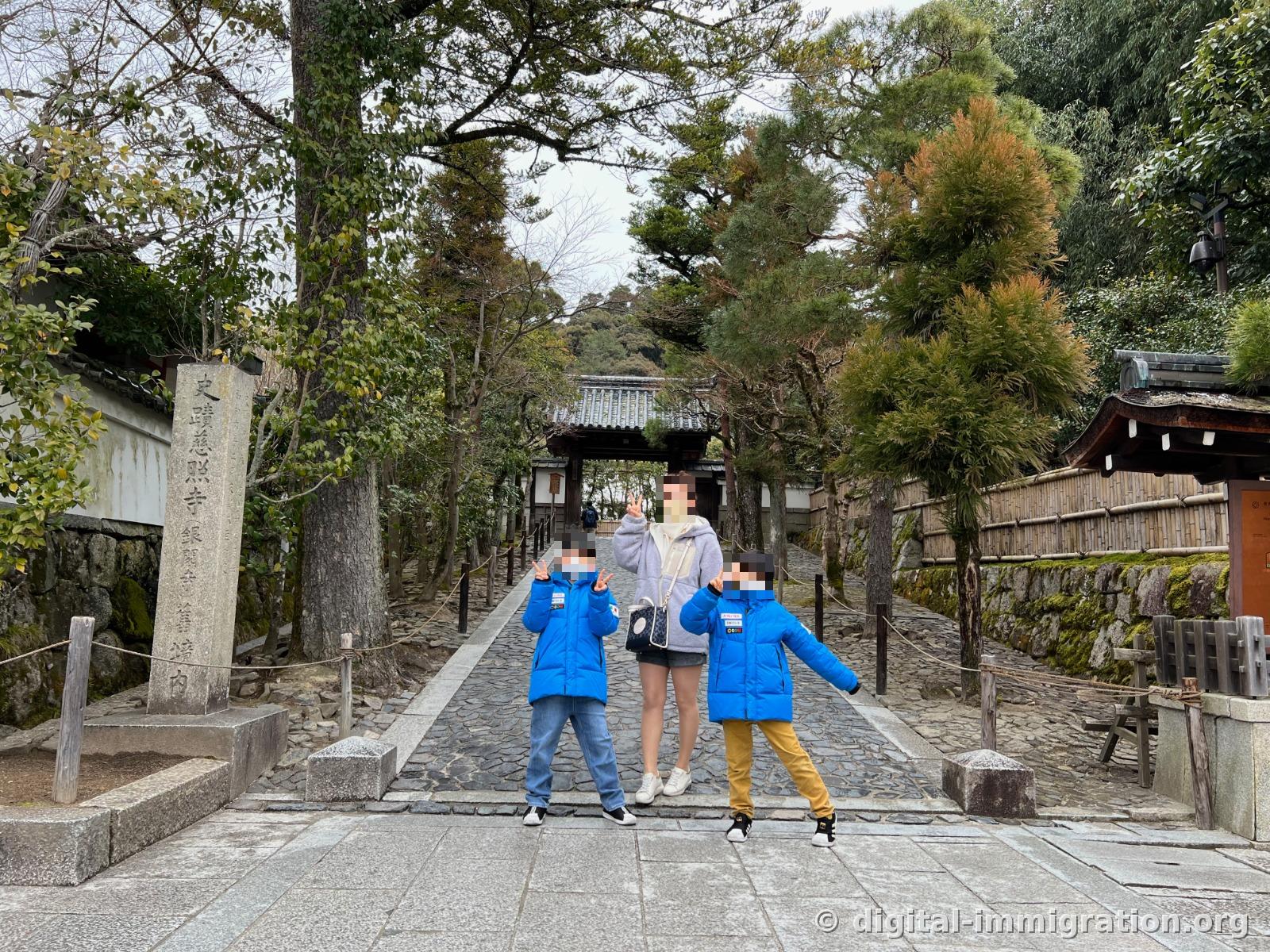 史蹟 慈照寺 銀閣寺 舊境内이라 새겨진 돌기둥 옆, 자갈 박힌 참배로에서 아이 둘과 어른이 손을 뻗고 포즈를 취한 모습. 뒤로 기와지붕 총문이 보인다
