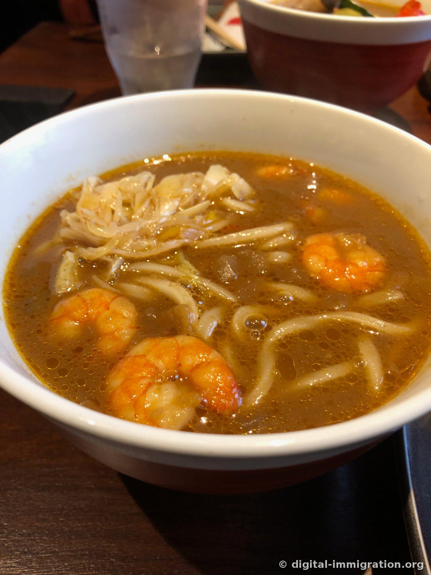 白い鉢に盛られた濃い茶色のカレースープうどん。殻付きえびと細切りした玉ねぎ、太いうどん麺が見える