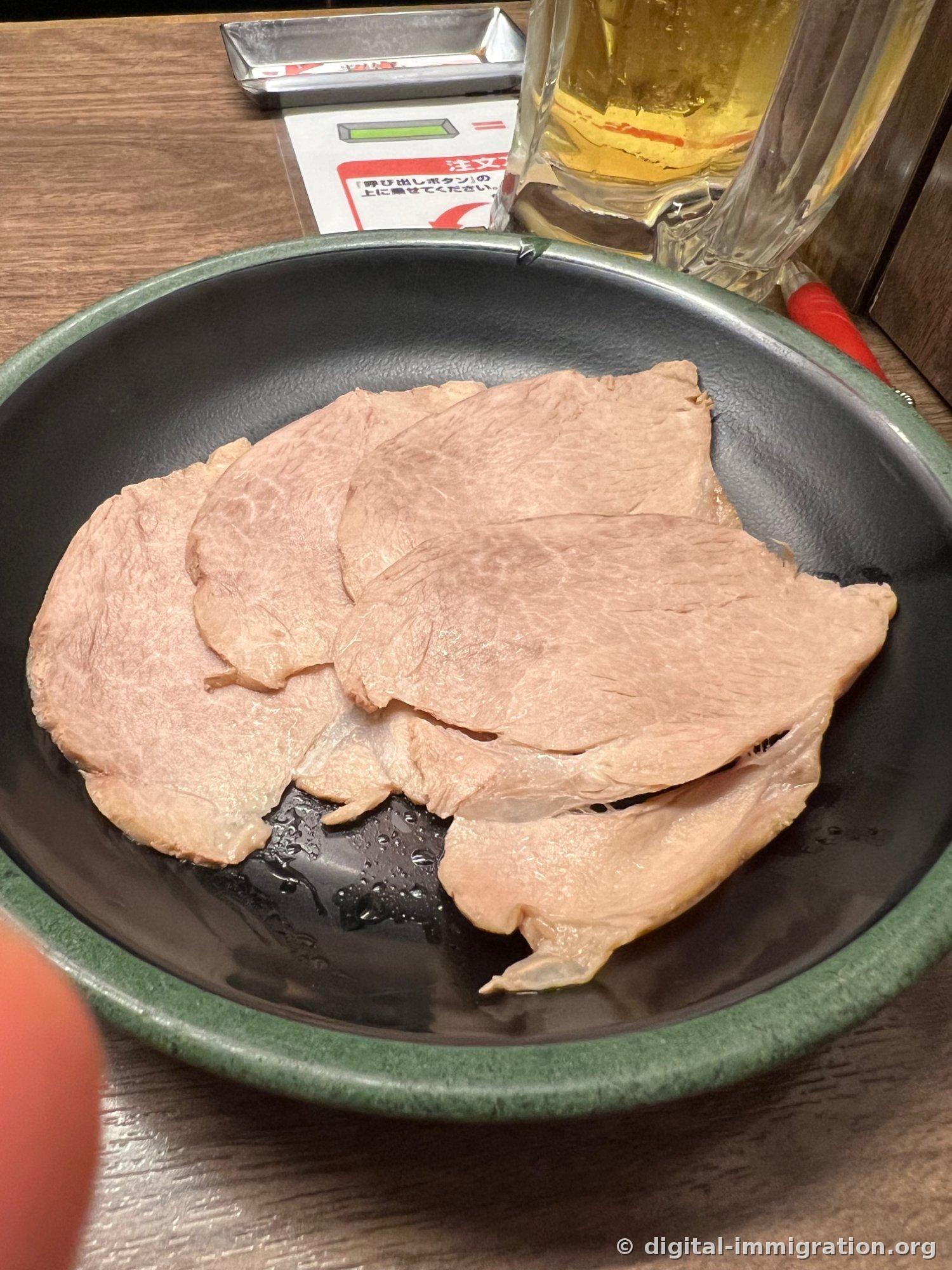 Multiple thinly sliced char siu pieces on a black plate with green border, with a draft beer glass, instruction sheet, and call button visible in the back