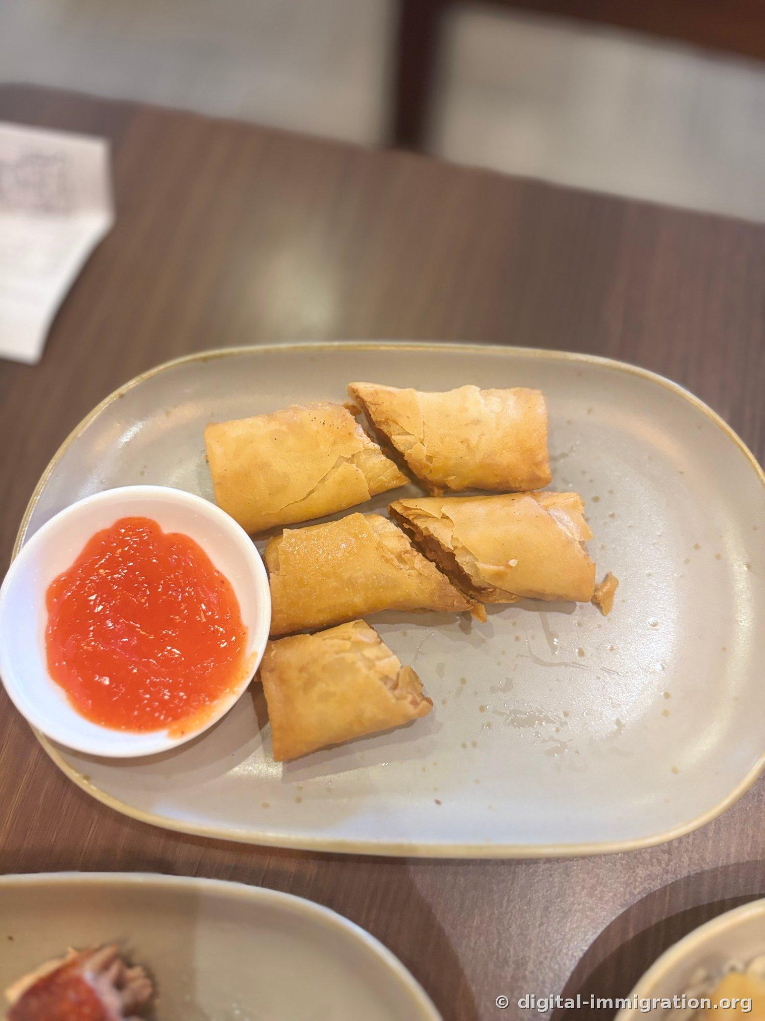 Five pieces of halved fried lumpia on a plate with a white small bowl of red chili sauce on the left