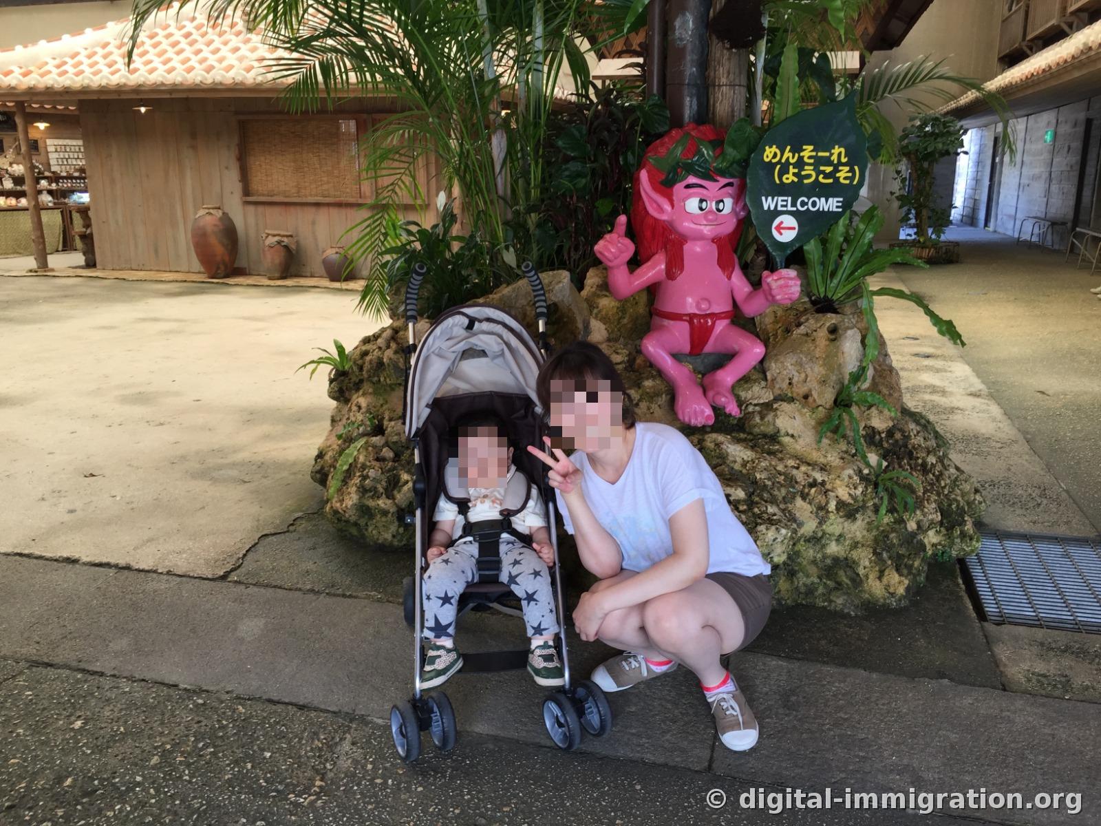 Devant la figurine de Kijimuna rose et le panneau « めんそーれ WELCOME », un visiteur est assis avec une poussette