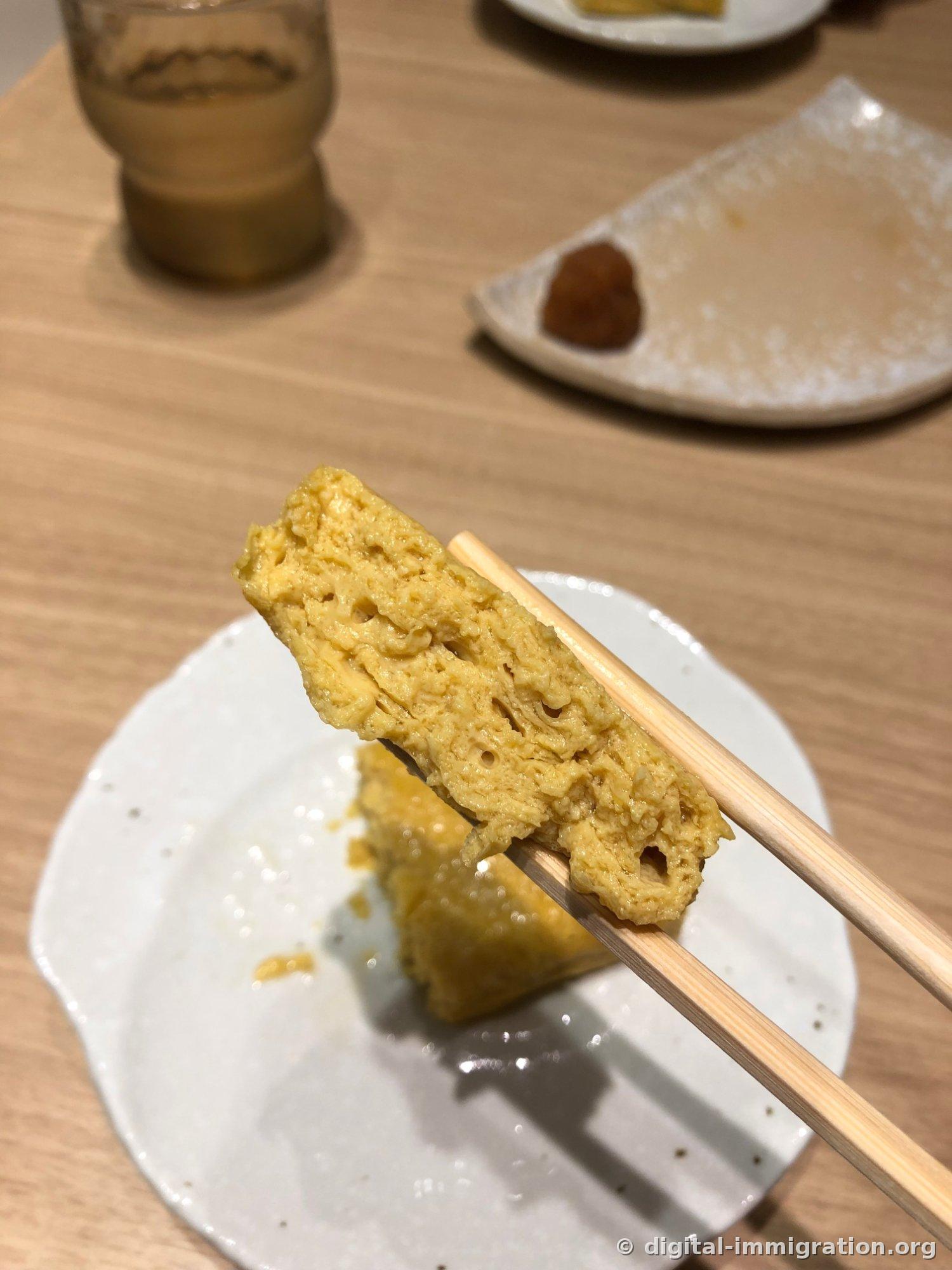 A tamagoyaki piece held with chopsticks showing the cross-section of layered egg with holes, with daikon remaining on the plate behind