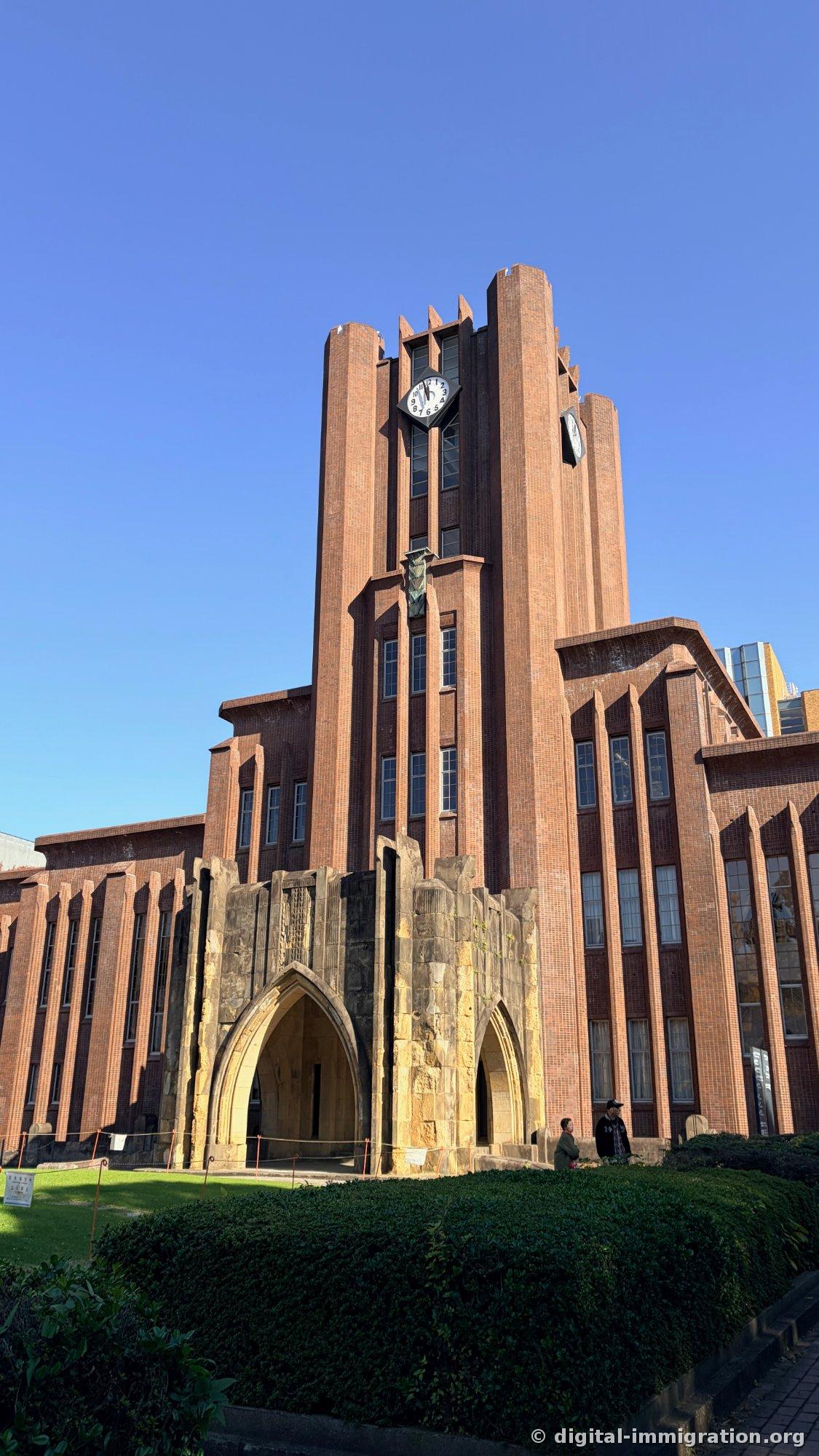 Yasuda Auditorium's clock tower viewed from below, with two octagonal clocks and a pointed arched entrance in rough stone, a trimmed green hedge in the foreground