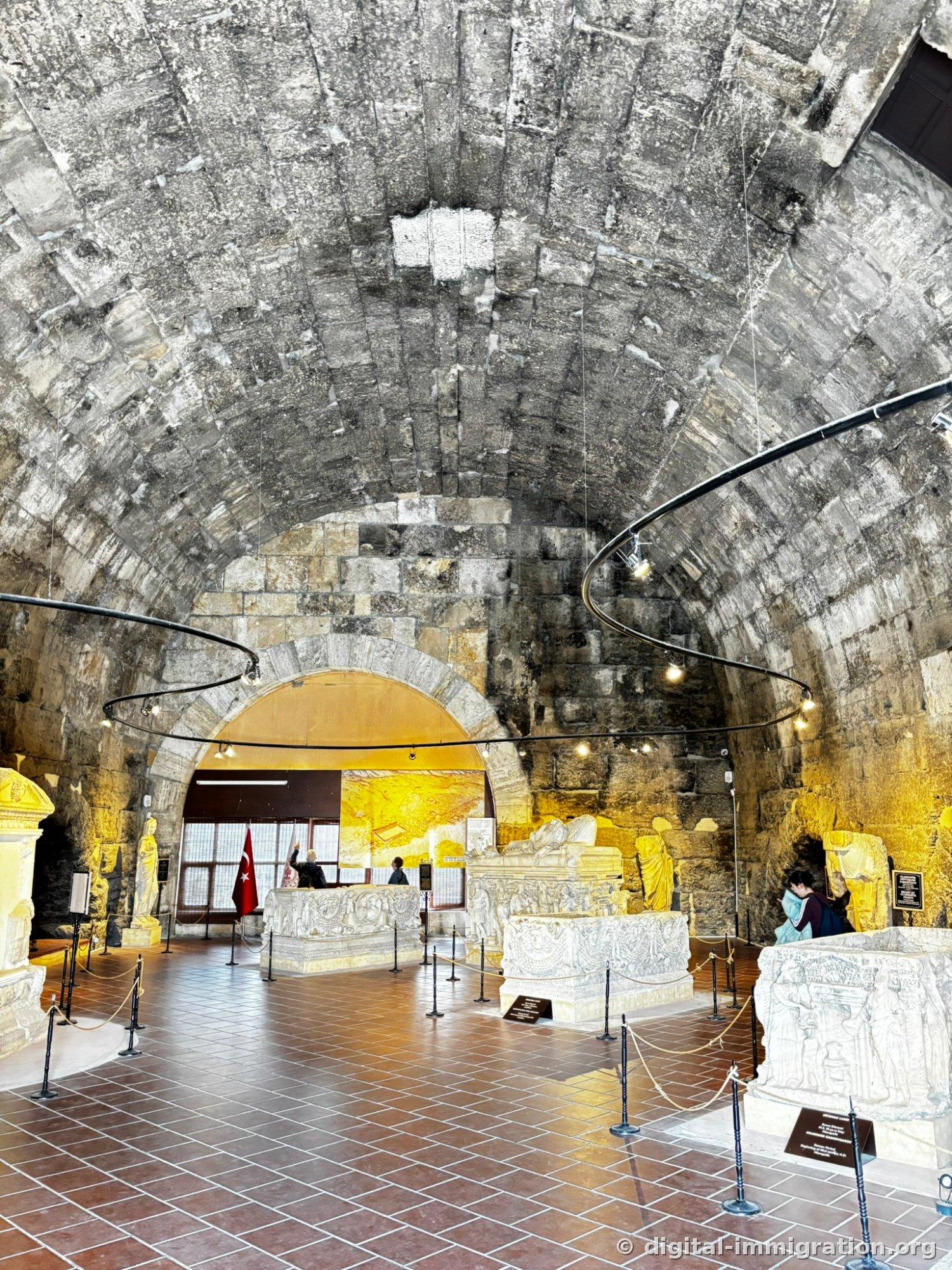 Interior of exhibition space with massive stone barrel vault ceiling, large sarcophagi displayed and cordoned off with rope on a red tile floor, Turkish flag hanging in the background