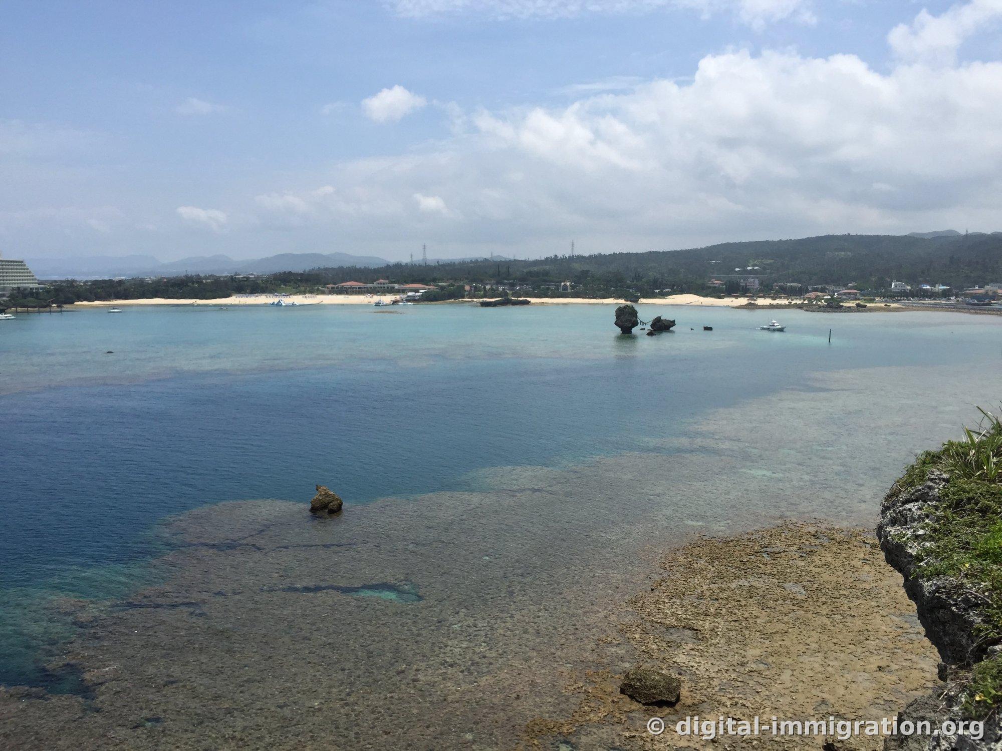 万座毛から眺めた恩納村の海岸線。白い砂浜と海浜施設、停泊したボートが見え、浅い海の上に小さな岩島がいくつか突き出ている