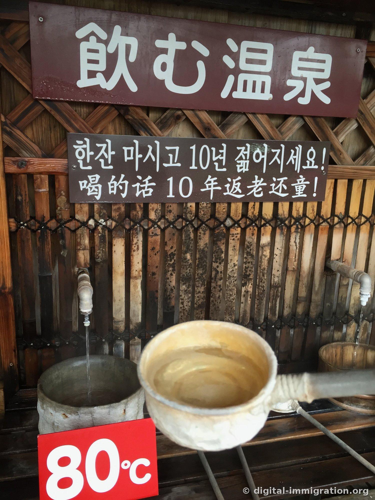 Une pancarte lisant '飲む温泉' et une affiche en coréen disant '한잔 마시고 10년 젊어지세요!' sous deux robinets avec de l'eau chaude coulant et une louche posée à côté, marquée 80℃