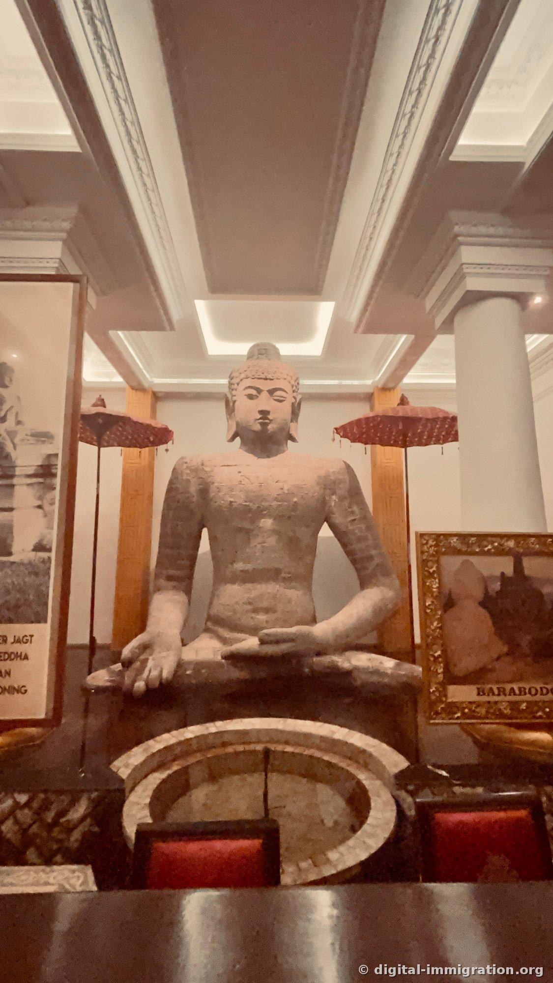Large Borobudur-style stone Buddha seated in lotus position in bright hall with white columns, red Balinese parasols on both sides