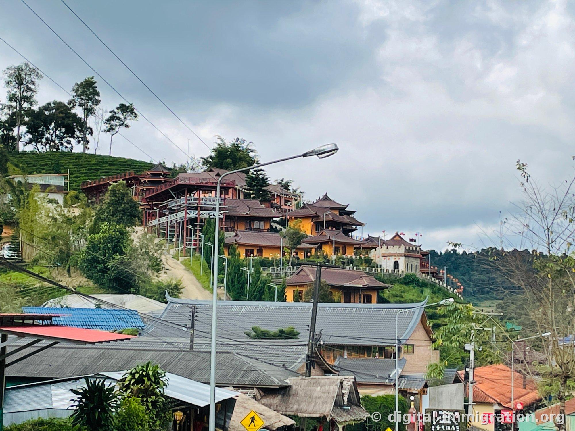 Vue du centre du village Ban Rak Thai depuis le bas. Au-dessus des maisons grises aux tuiles, on voit des bâtiments aux murs jaunes avec des avant-toits relevés, des lanternes rouges, et une structure blanche en forme de remparts