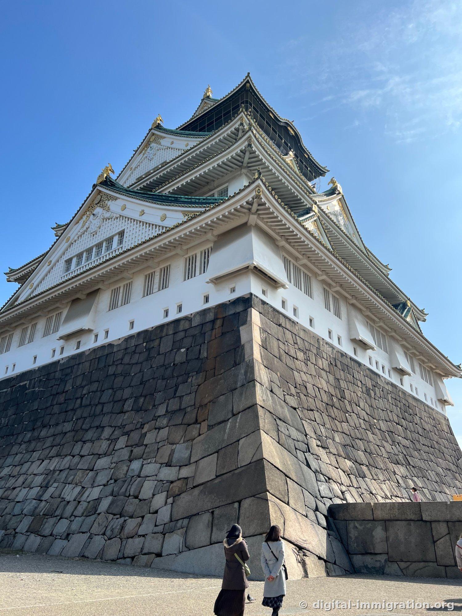 Vue de bas en haut de la tour de guet du château d'Osaka. L'énorme mur de pierre noir avec ses murs blancs et toit orné d'or qui s'élèvent en couches, et deux visiteurs debout en bas