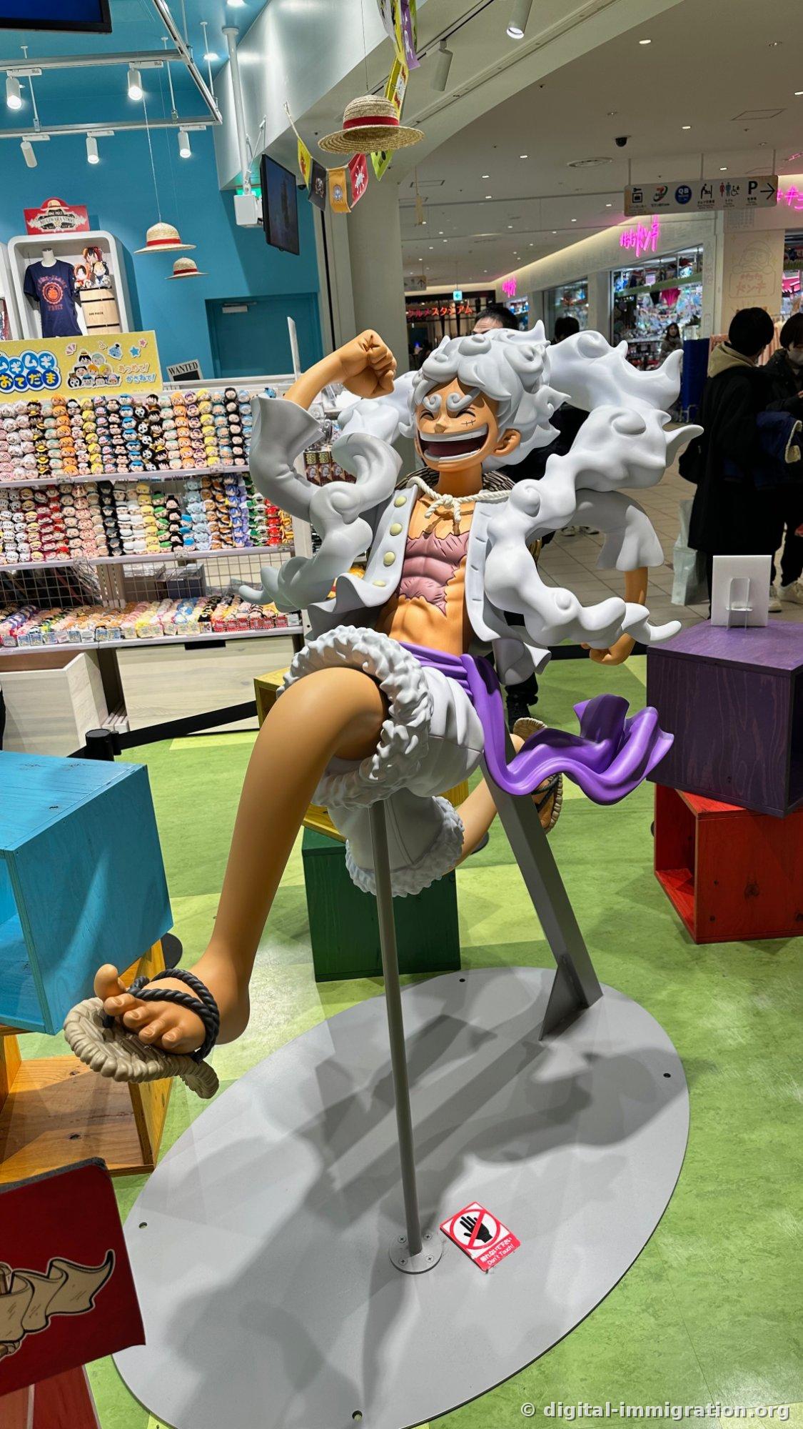 Inside a shopping mall One Piece store, a life-size Gear 5 Luffy figure sitting and smiling on a green floor, straw hats and character merchandise displays on sky-blue walls in the background
