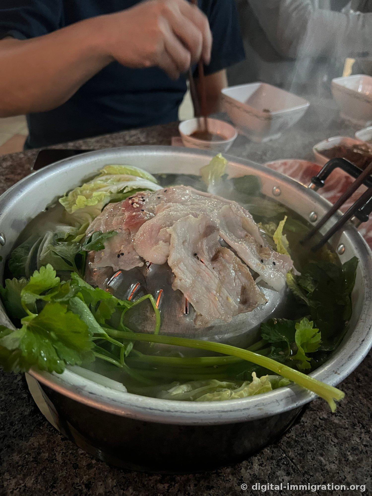 Pork belly cooking on the dome of the mookata grill, with cabbage, cilantro, and water dropwort submerged in broth in the edge groove, steam rising