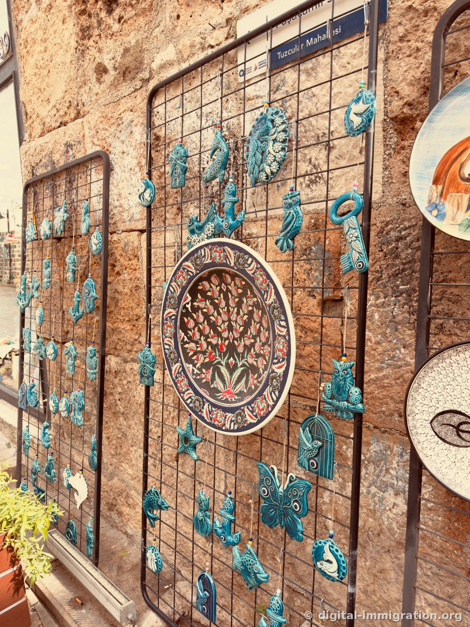 Étalage en treillis accrochés à un mur de pierre avec des décorations en céramique bleu-vert (hiboux, papillons, poissons, clés, mains). Au centre, un grand plat aux motifs de tulipes. Au-dessus, le panneau Tuzcular Mahallesi