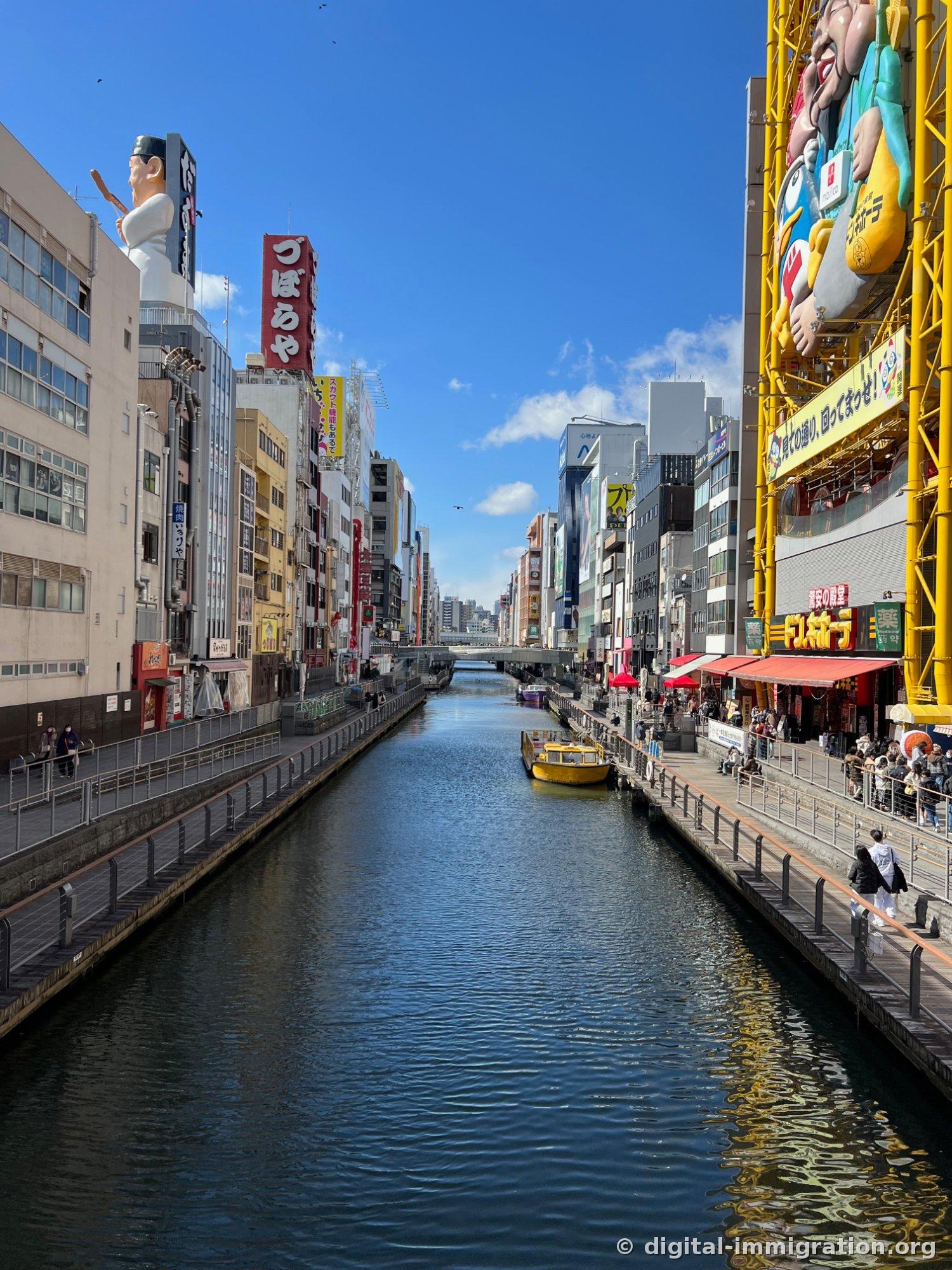 Vue de jour du Dotonbori, fleuve clair avec l'enseigne Fugu Zuboraya et la poupée Kuidaore de chaque côté, la grande roue du Don Quixote, et un bateau de croisière jaune amarré