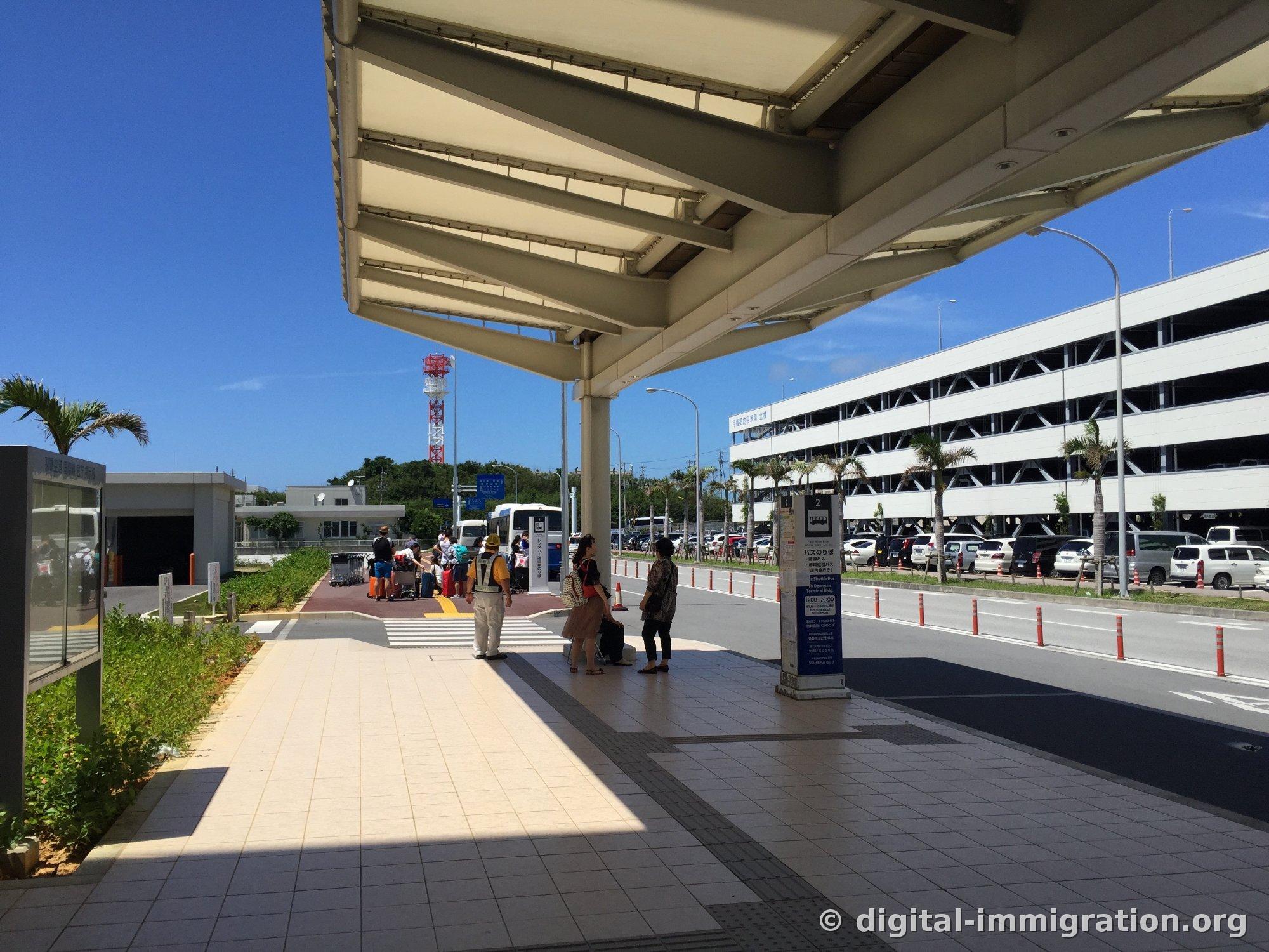 Arrêt de bus à l'extérieur du terminal de l'aéroport de Naha. Des personnes avec des bagages sont debout sous le dais, avec des palmiers, un immeuble de stationnement à plusieurs étages et une tour en rouge et blanc sous le ciel bleu.