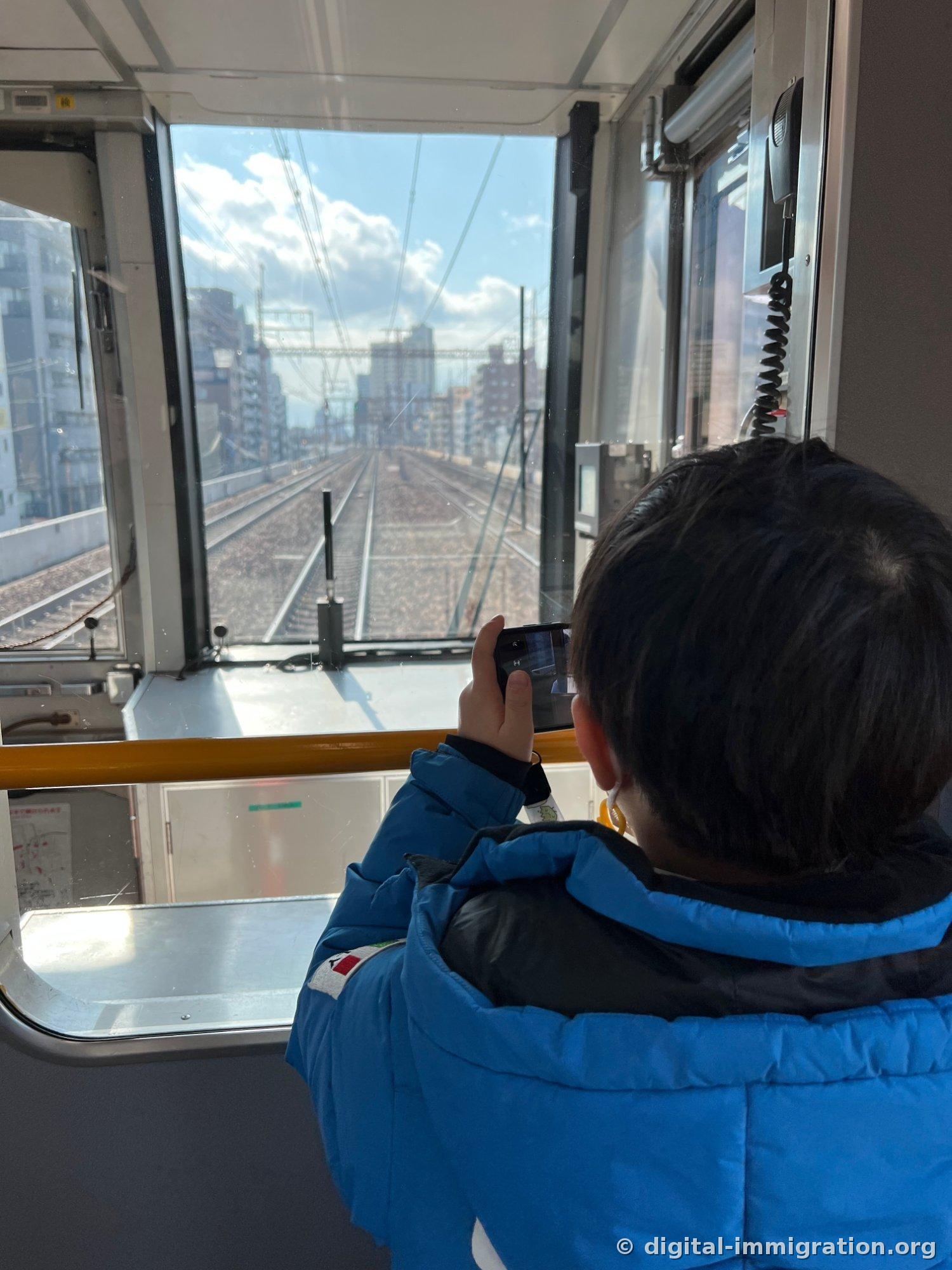 Un enfant en doudoune bleue collé contre la vitre derrière la cabine du conducteur à l'avant du train, photographiant les rails avec son téléphone