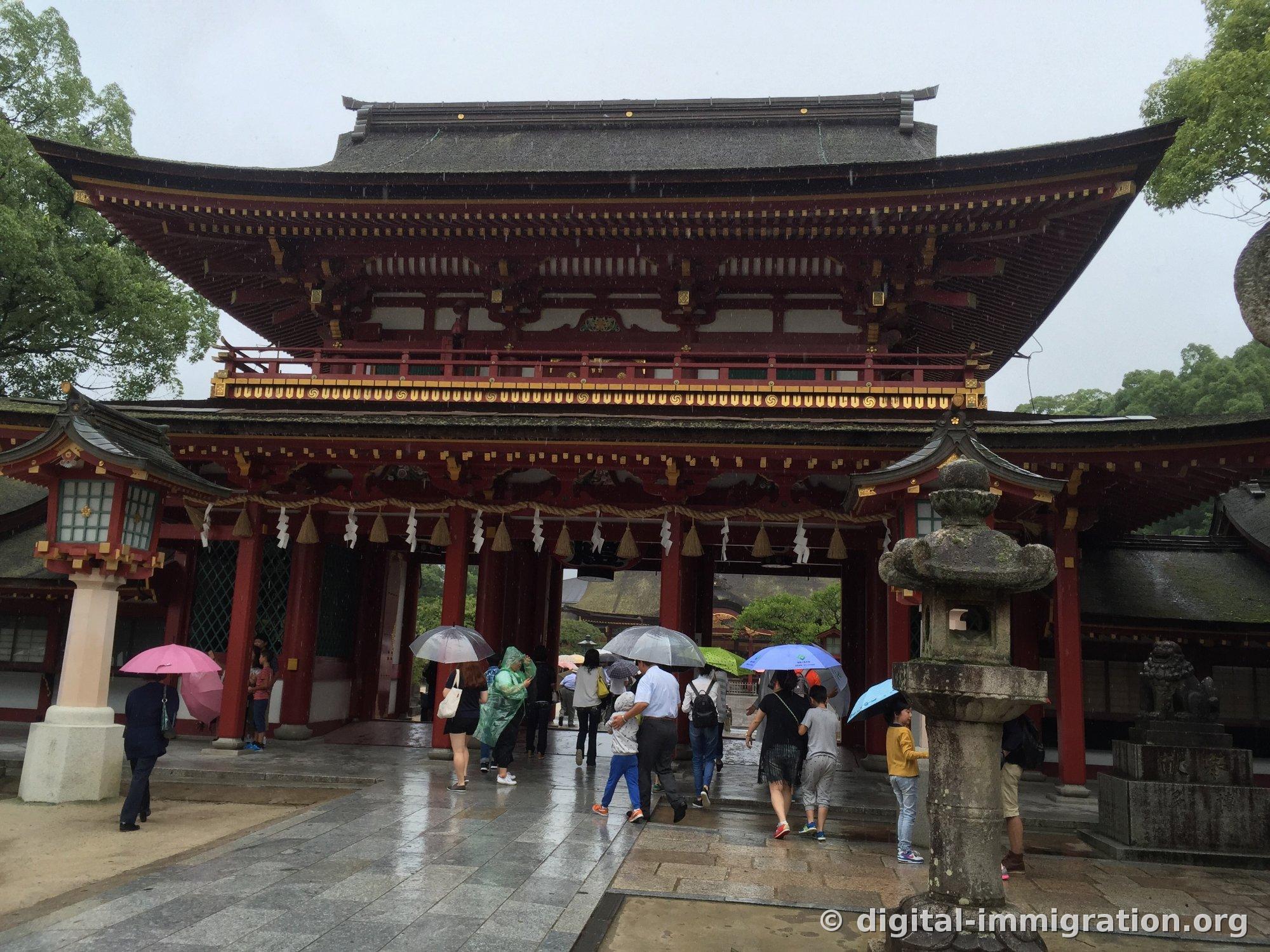 Front of two-story crimson romon gate with sacred rope and white shide under the eaves, worshippers with umbrellas passing through