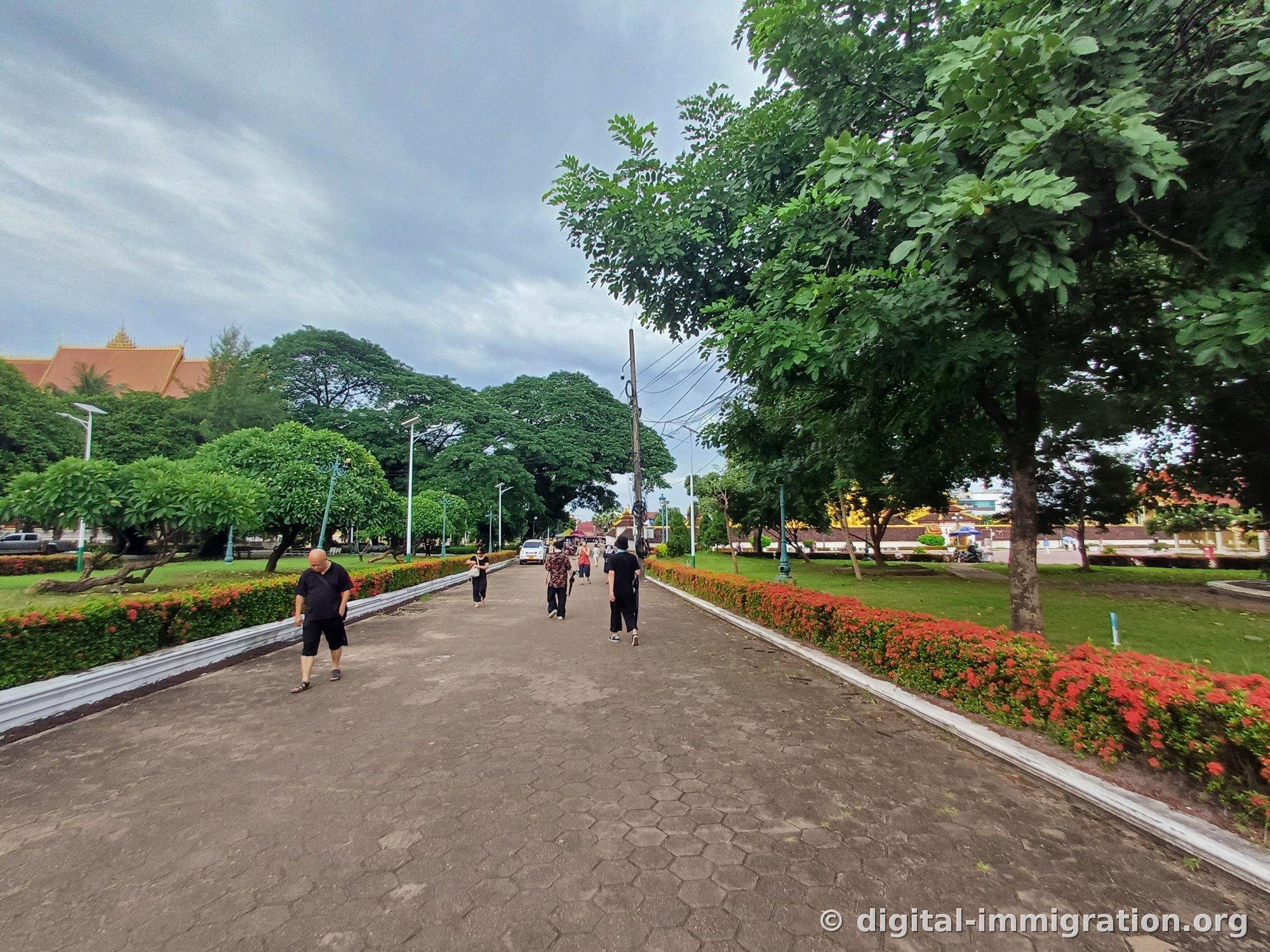 Shaded promenade section with local people walking, the golden stupa roof visible in the distance beyond the wall