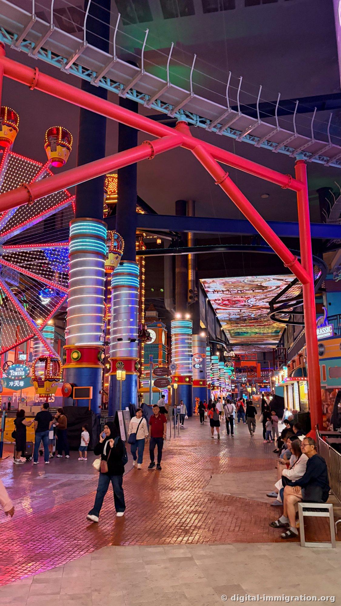 Indoor theme park interior with red steel and neon pillars, SKY TOWERS sign and Ferris wheel, people walking below large LED screen ceiling