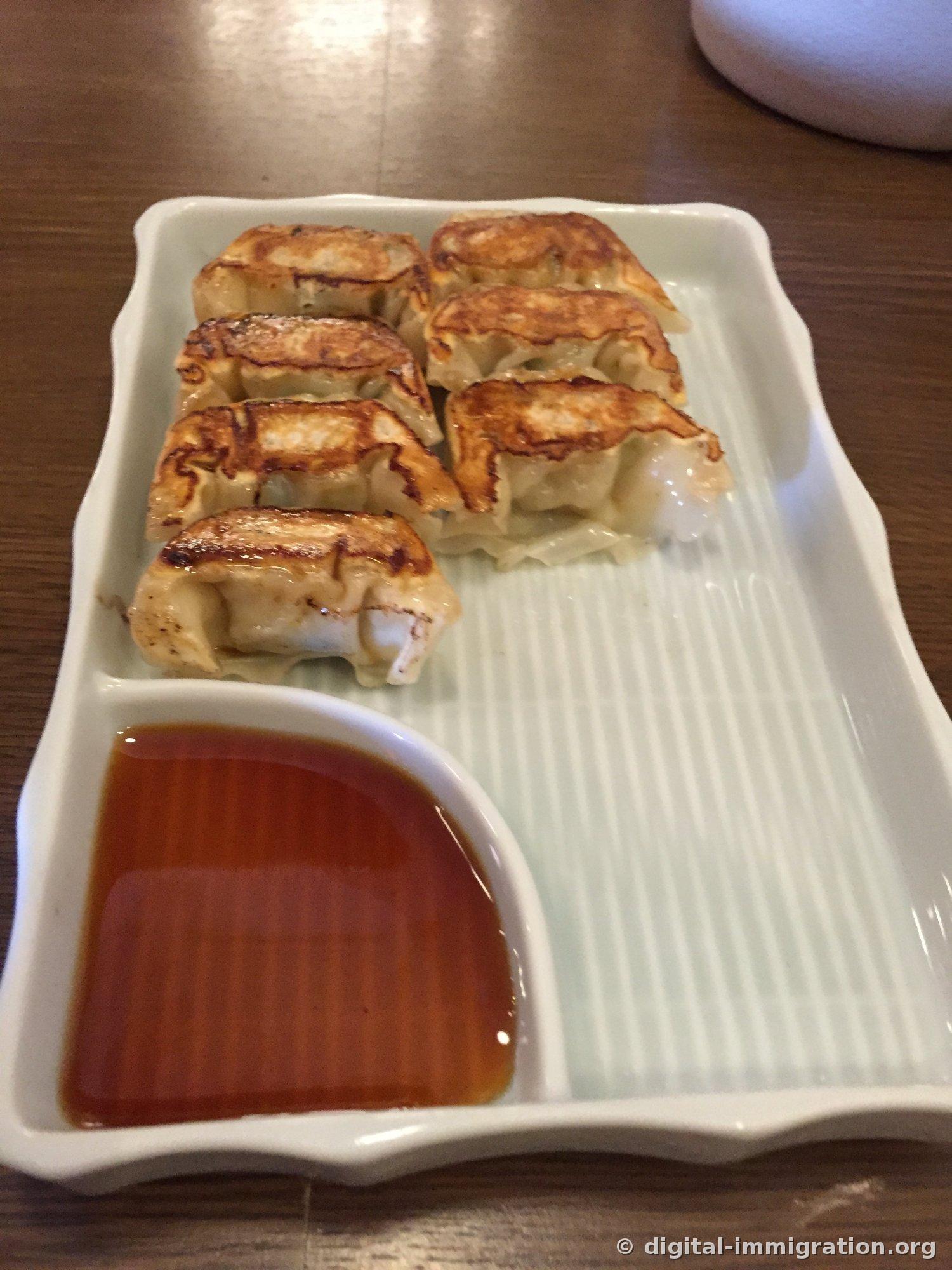 Sept gyoza Hakata Hitokhuchi dorés et grillés alignés sur une assiette blanche rectangulaire, sauce soja rougeâtre servie dans le compartiment de gauche
