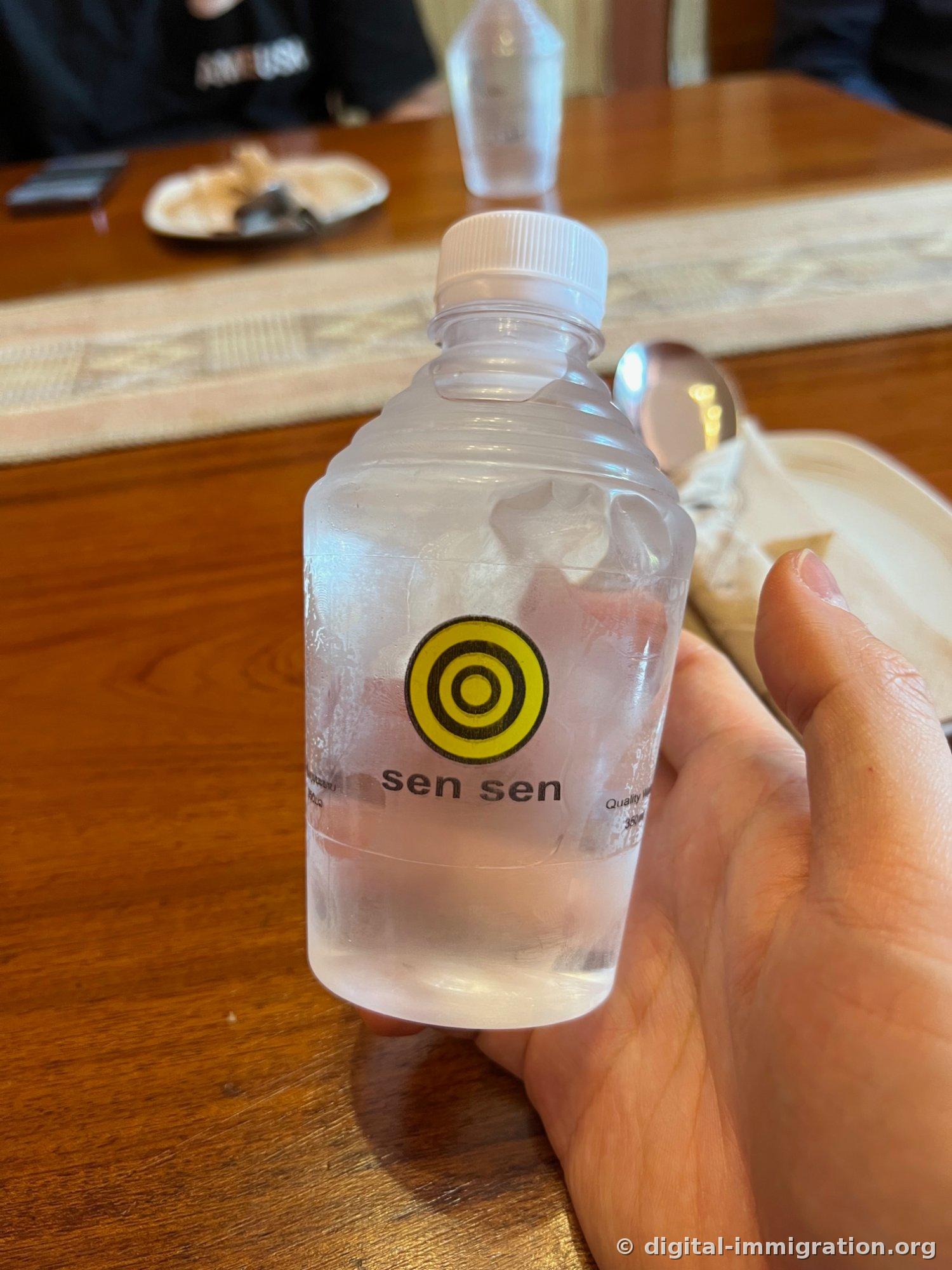 노란 동심원 로고와 'sen sen' 글자가 인쇄된 350ml 생수병을 손에 쥔 모습, 뒤로 원목 테이블과 러너