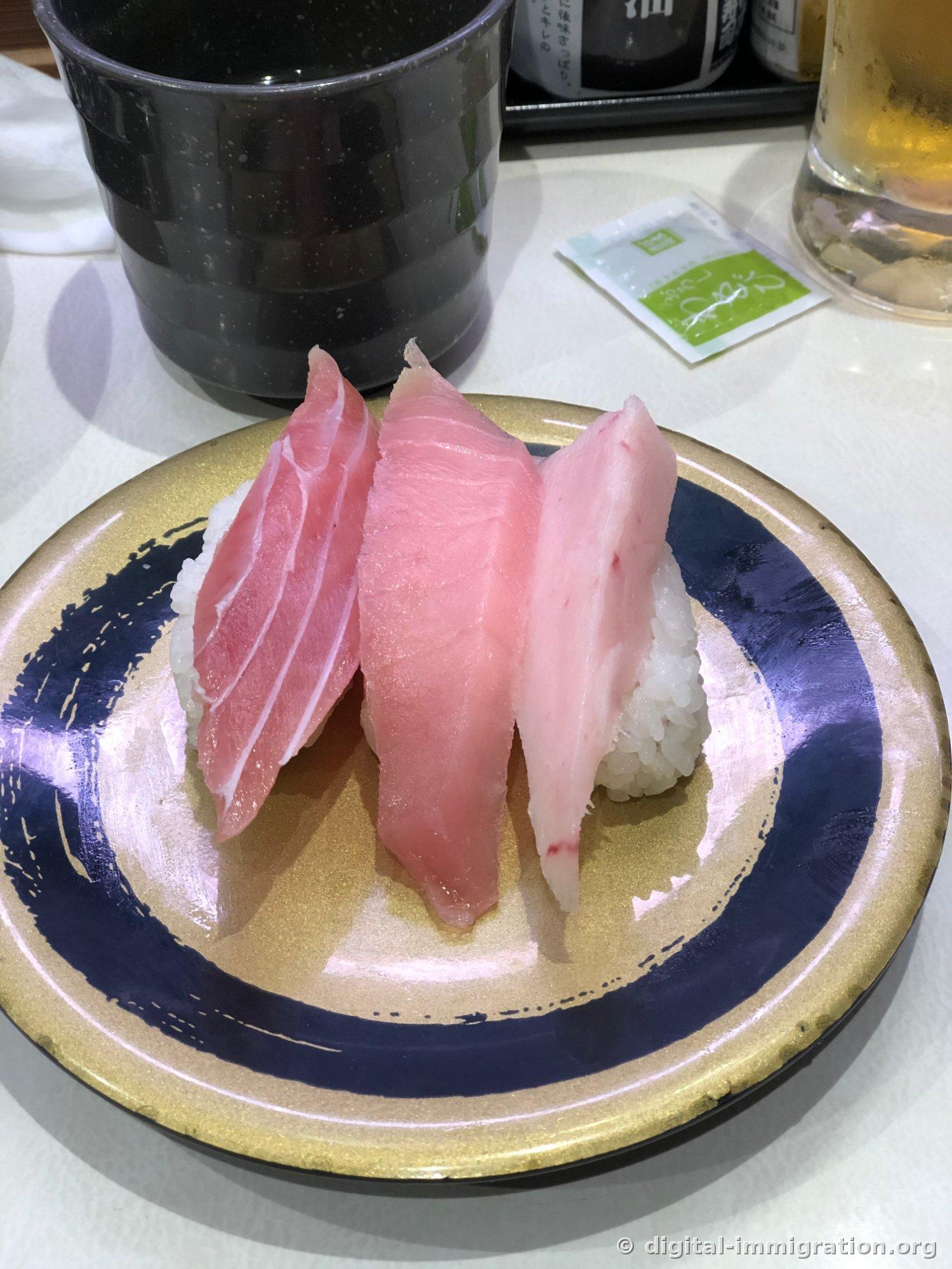 Sur une assiette avec des bordures dorées et bleu marine, trois types de sushi au thon - viande rouge, thon aileron et ventre - sont disposés côte à côte, avec une tasse de thé noire et un verre de bière à l'arrière