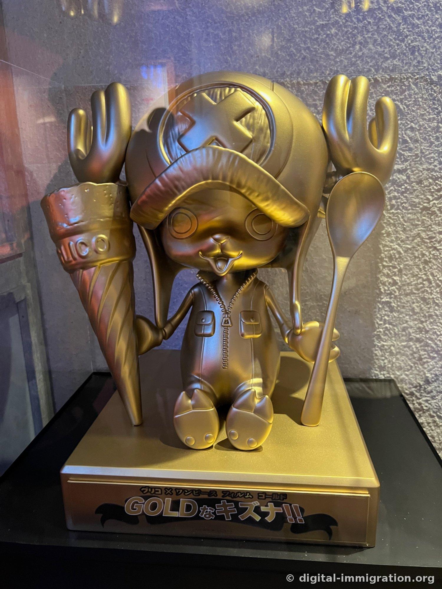 A golden Chopper figure in a glass display case with 'Glico × One Piece Film Gold' written on the base