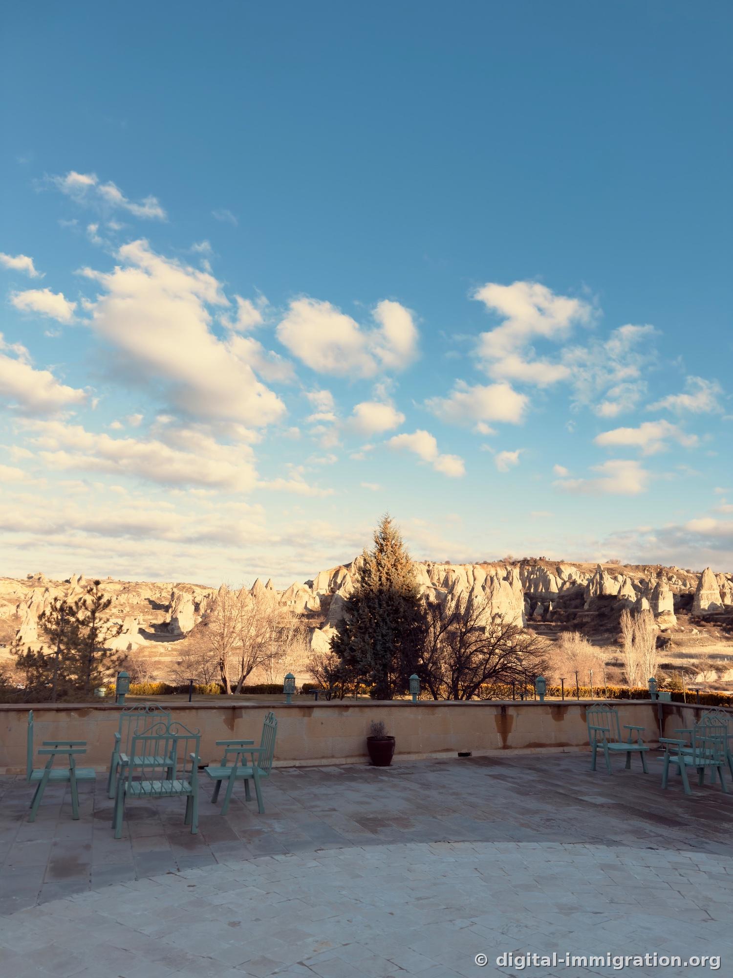 Un ciel couvert de petits nuages, une large terrasse en pierre pavée et des chaises menthe, au premier plan la crête des cheminées de fée de Göreme et des arbres aux feuilles tombées